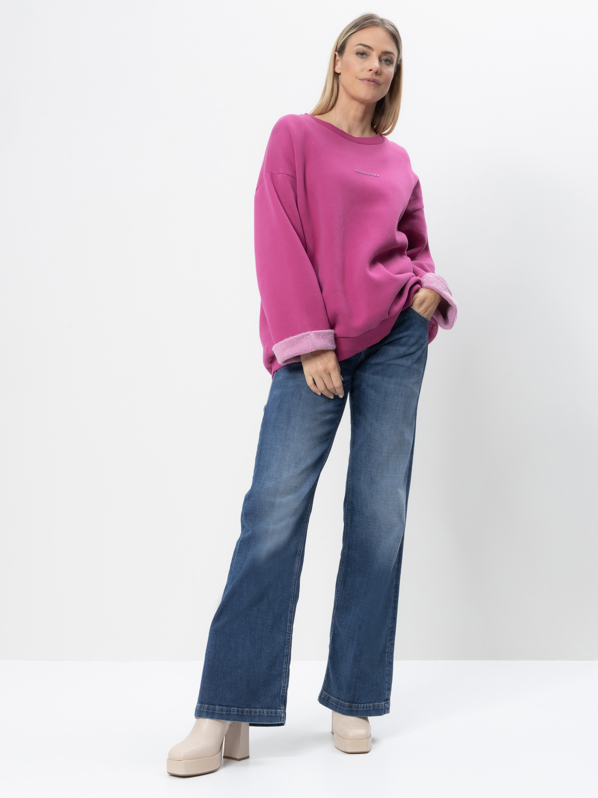 Eine Frau steht vor einem weißen Hintergrund und trägt ein lockeres rosafarbenes Sweatshirt mit hochgekrempelten Ärmeln, weit geschnittene Bluejeans und beigefarbene Plateauschuhe. Sie hat glattes, schulterlanges blondes Haar und einen entspannten Gesi