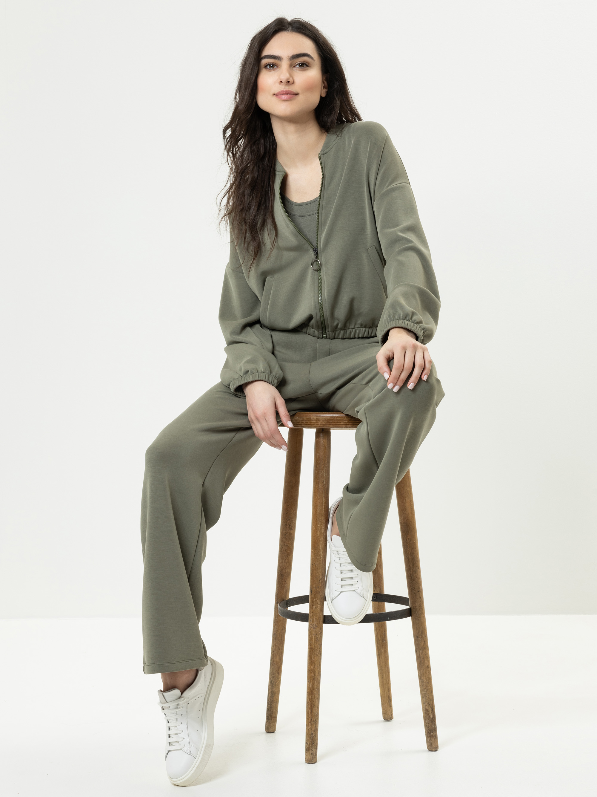 Eine Frau mit langen dunklen Haaren sitzt auf einem Holzschemel und trägt ein passendes olivgrünes Outfit mit Jacke und Hose sowie weiße Turnschuhe vor einem schlichten weißen Hintergrund.