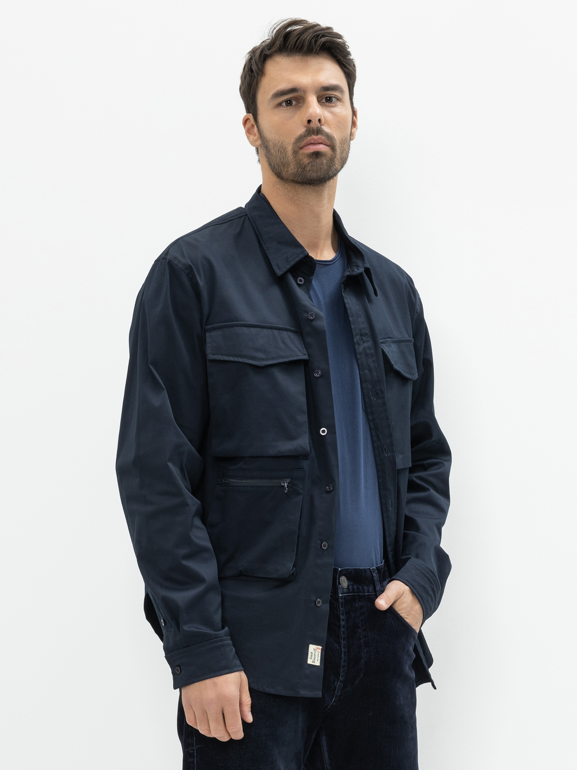 Ein Mann mit kurzen braunen Haaren und einem Bart trägt eine marineblaue Button-up-Jacke über einem passenden Hemd und eine dunkle Hose. Er steht vor einem schlichten weißen Hintergrund und hat eine Hand in der Tasche.