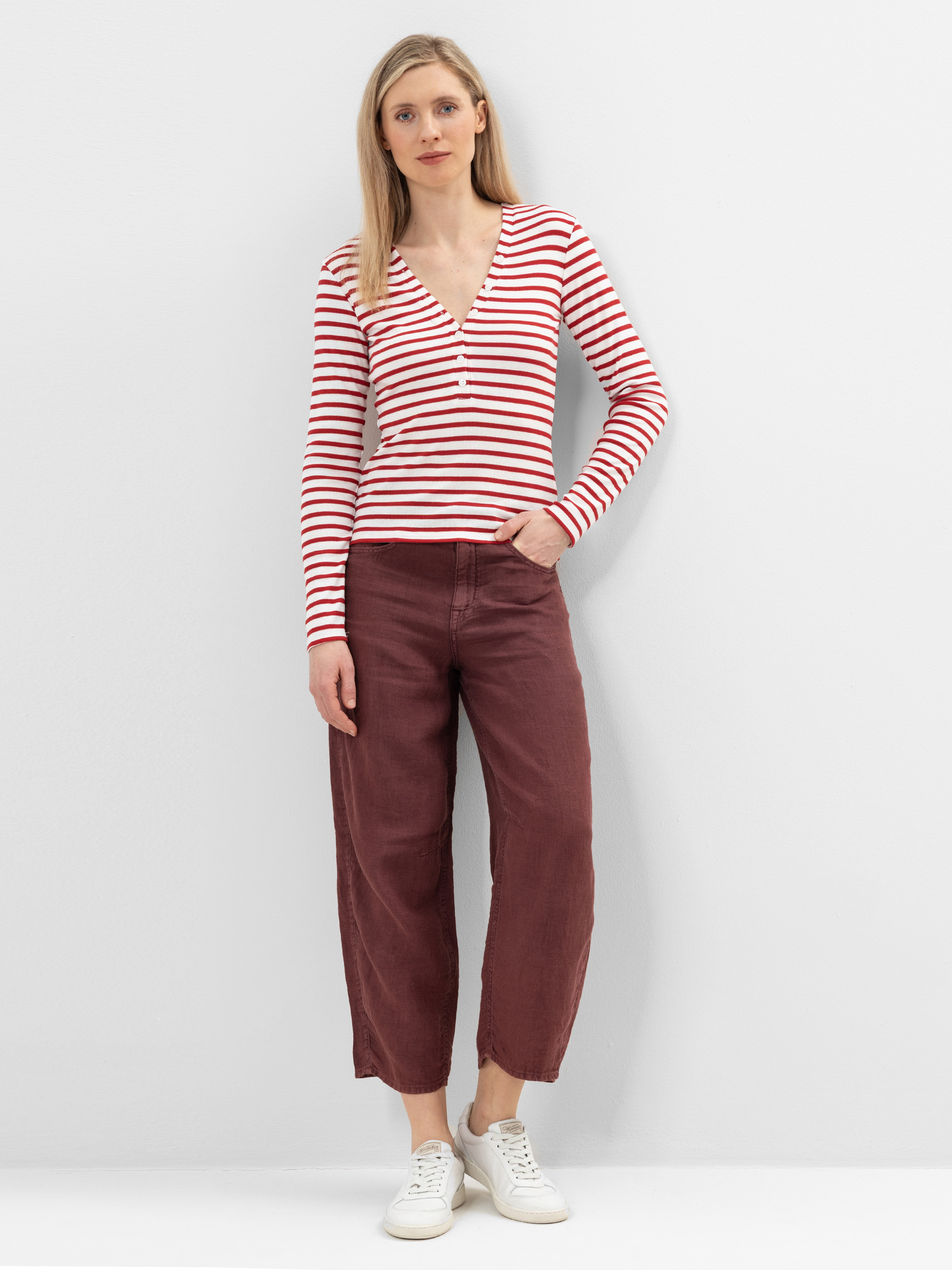 Eine Frau steht vor einer schlichten weißen Wand. Sie trägt ein langärmeliges, rot-weiß gestreiftes Oberteil, eine kastanienbraune, weit geschnittene Hose und weiße Turnschuhe. Sie hat glattes blondes Haar und einen neutralen Gesichtsausdruck.