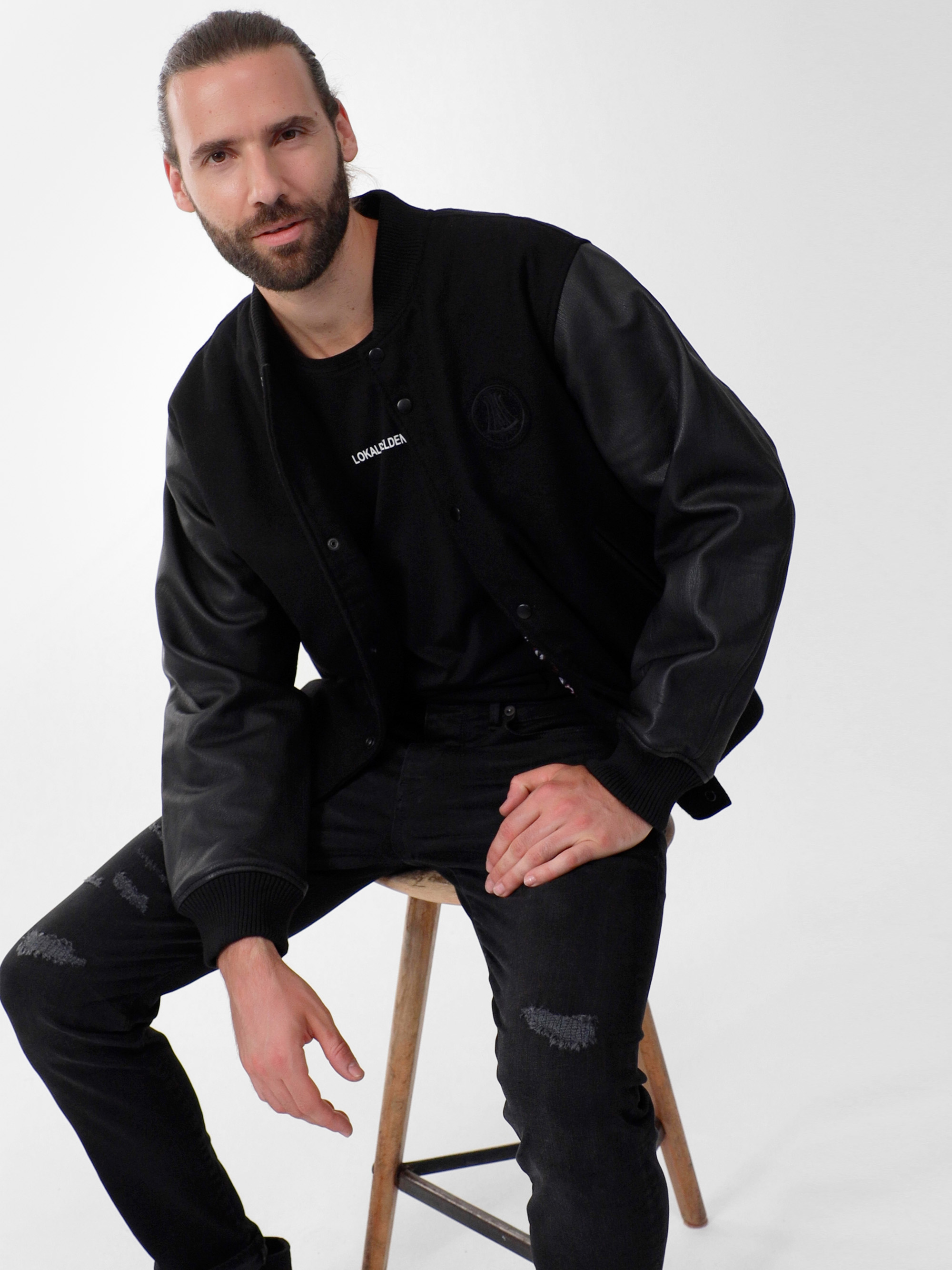 Ein Mann mit langen, zurückgebundenen Haaren und Bart sitzt auf einem Holzschemel und trägt eine schwarze Sportjacke mit Lederärmeln, ein schwarzes T-Shirt und eine zerrissene schwarze Jeans vor einem schlichten weißen Hintergrund.