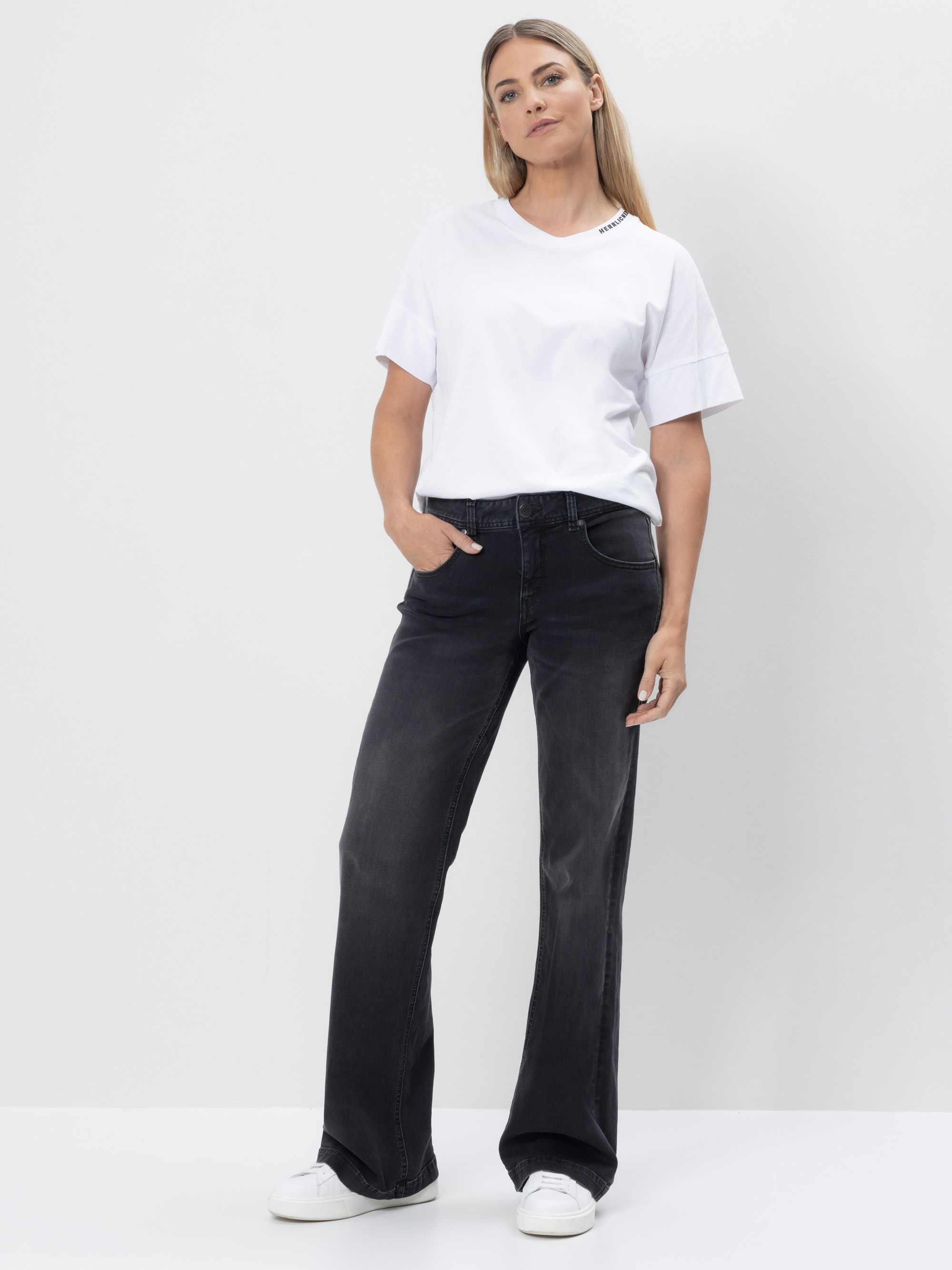 Eine Frau steht vor einem weißen Hintergrund und trägt ein weißes T-Shirt, schwarze Jeans mit weitem Bein und weiße Turnschuhe. Sie hat langes, glattes Haar, eine Hand in der Hosentasche und schaut ruhig in die Kamera.