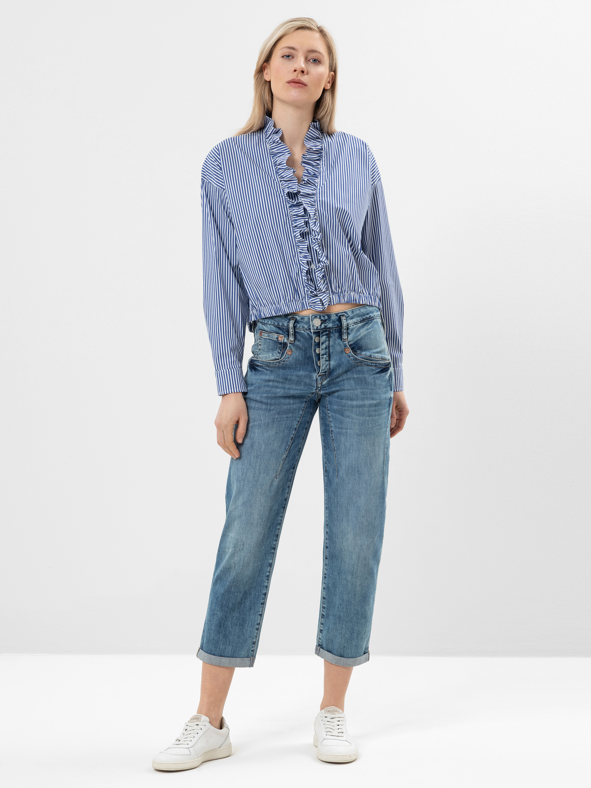 Eine Frau steht in einer entspannten Pose und trägt eine blau-weiß gestreifte Rüschenbluse, blaue Jeans und weiße Turnschuhe vor einem schlichten weißen Hintergrund.
