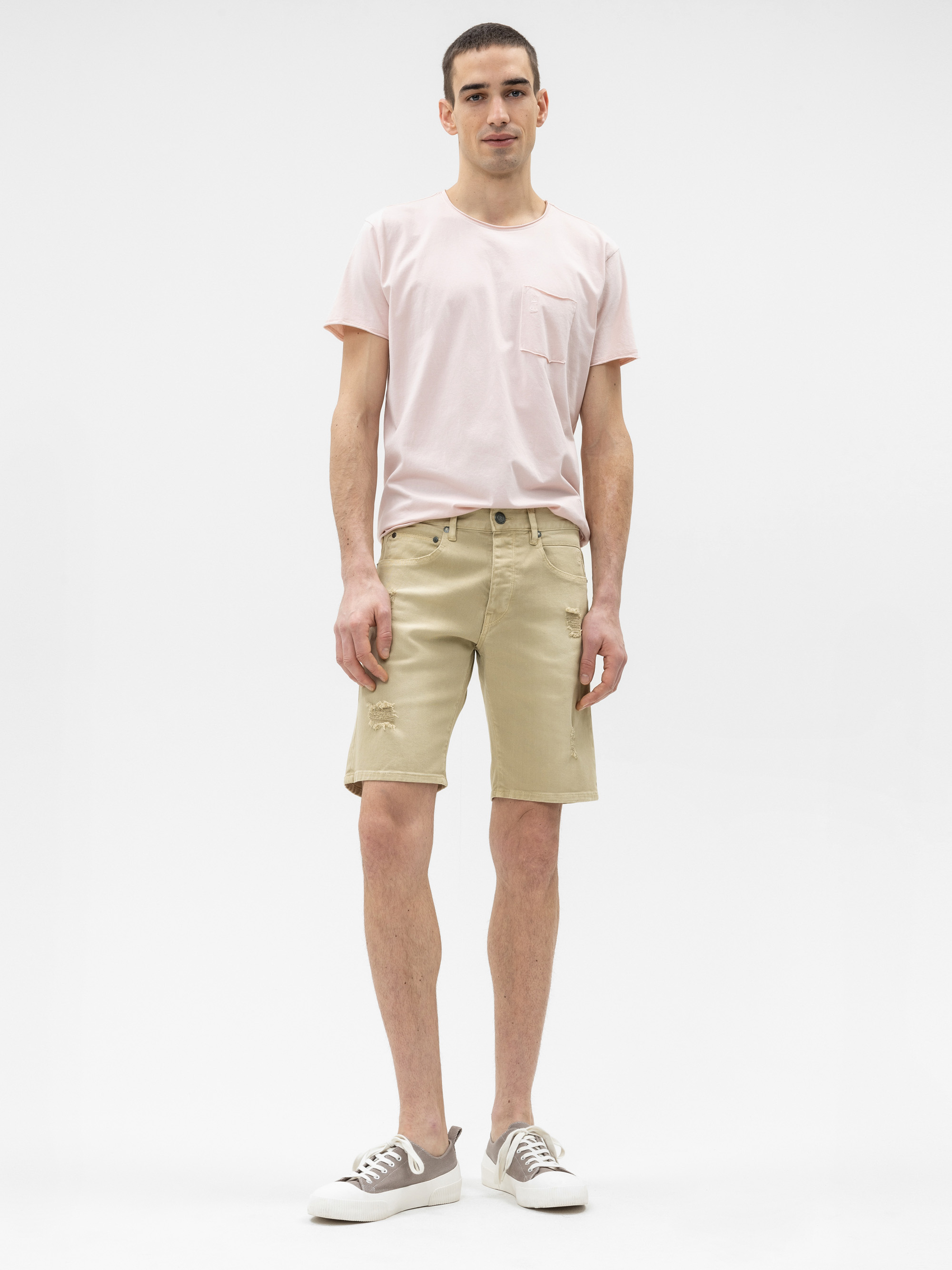 Ein Mann steht vor einem schlichten weißen Hintergrund und trägt ein hellrosa T-Shirt, beigefarbene knielange Shorts und graue Turnschuhe mit weißen Sohlen. Er blickt nach vorne und hat eine entspannte Miene und Haltung.