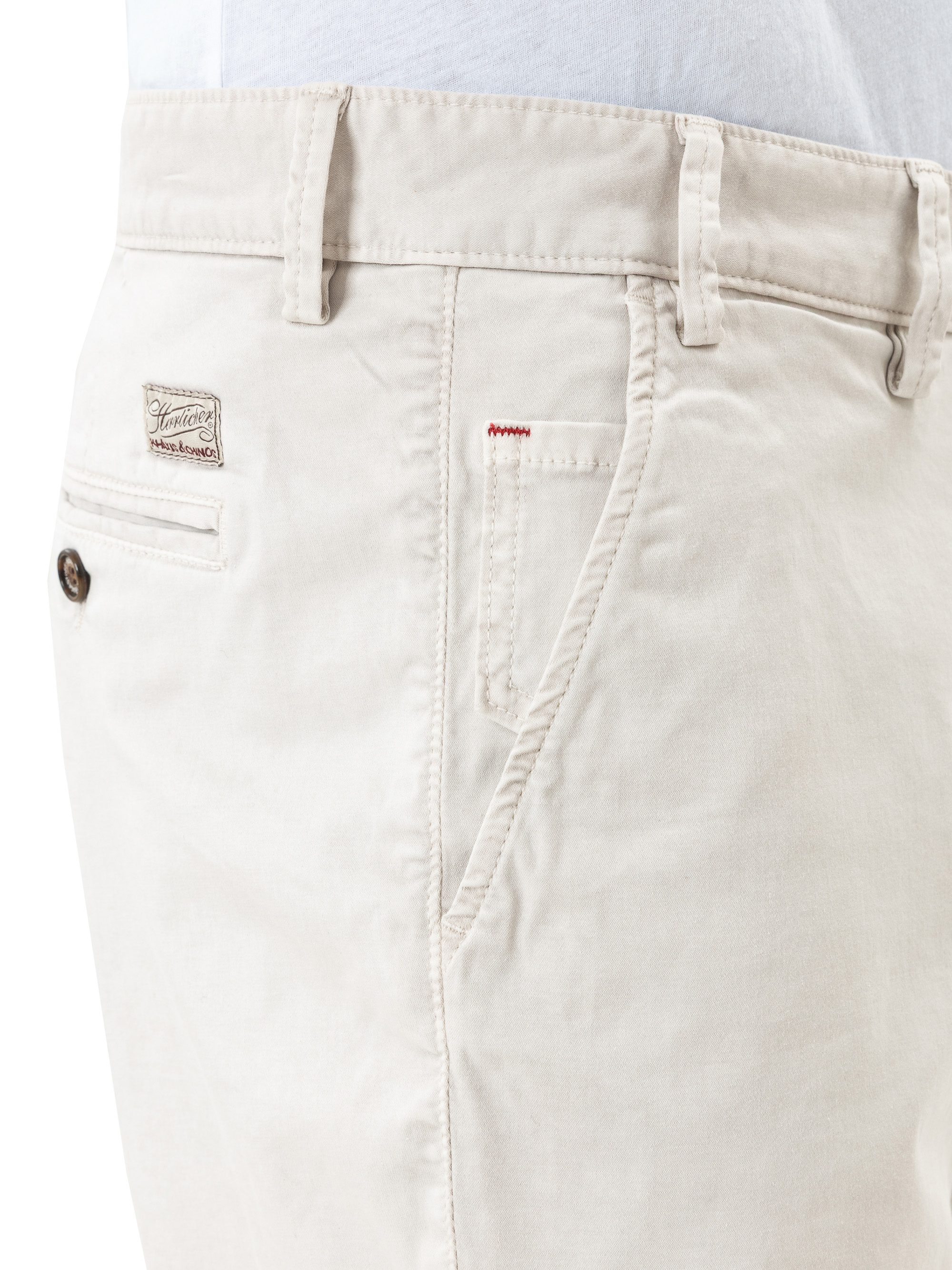 Nahaufnahme einer Person in einer hellbeigen Hose mit Gürtelschlaufen, einer Vordertasche mit roten Nähten und einer Gesäßtasche mit einem kleinen Logoaufnäher und einem braunen Knopf. Die Person trägt außerdem ein weißes Hemd, das in die Hose ges