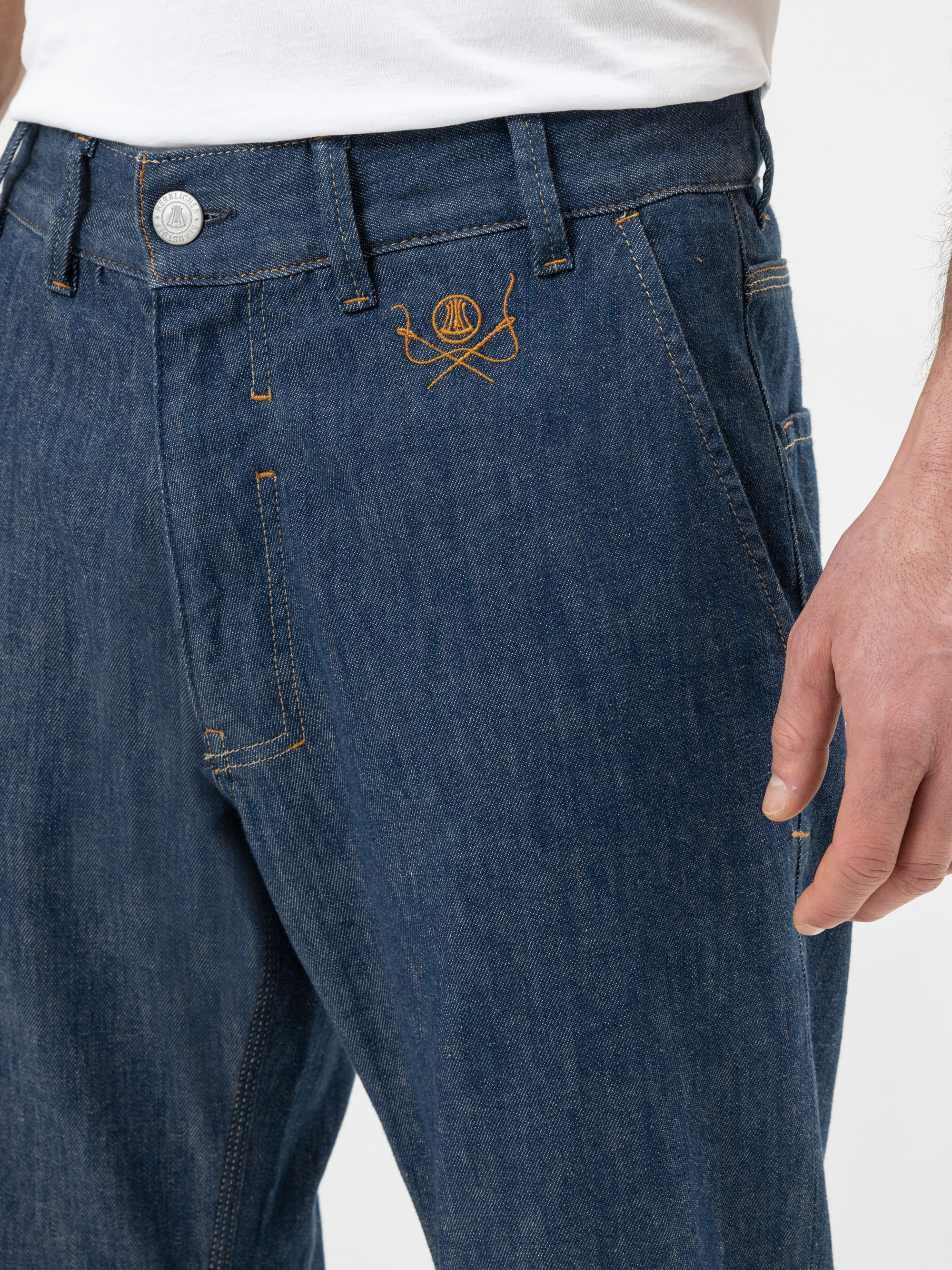 Eine Person trägt eine blaue Jeans und ein weißes Hemd. Die Jeans hat orangefarbene Nähte und ein aufgesticktes gelbes Logo in der Nähe der linken Vordertasche. Nur der untere Teil des Oberkörpers und die rechte Hand sind sichtbar.