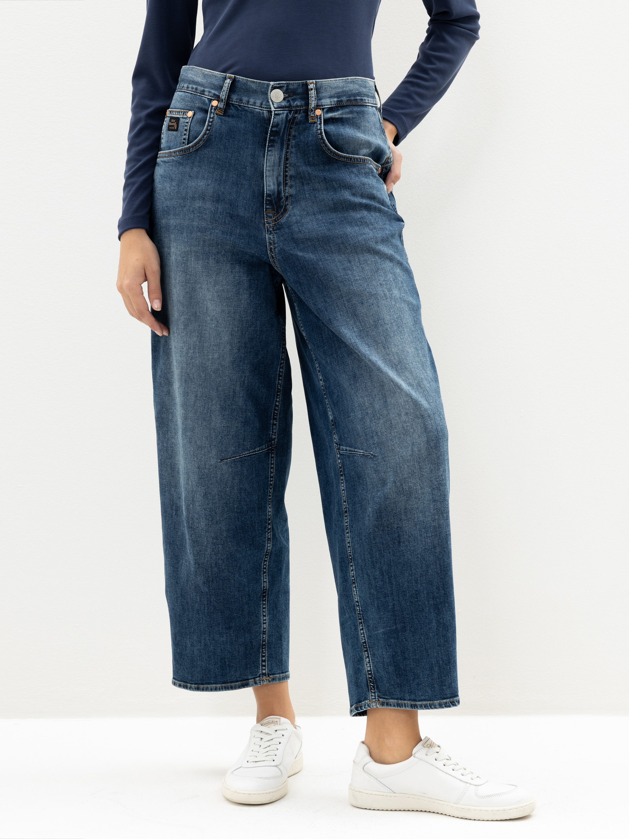Eine Person, die weit geschnittene blaue Jeans, ein langärmeliges marineblaues Oberteil und weiße Turnschuhe trägt, steht vor einem schlichten, hellen Hintergrund. Ihre linke Hand steckt in einer Tasche, und ihr Kopf ist auf dem Bild nicht zu sehen.