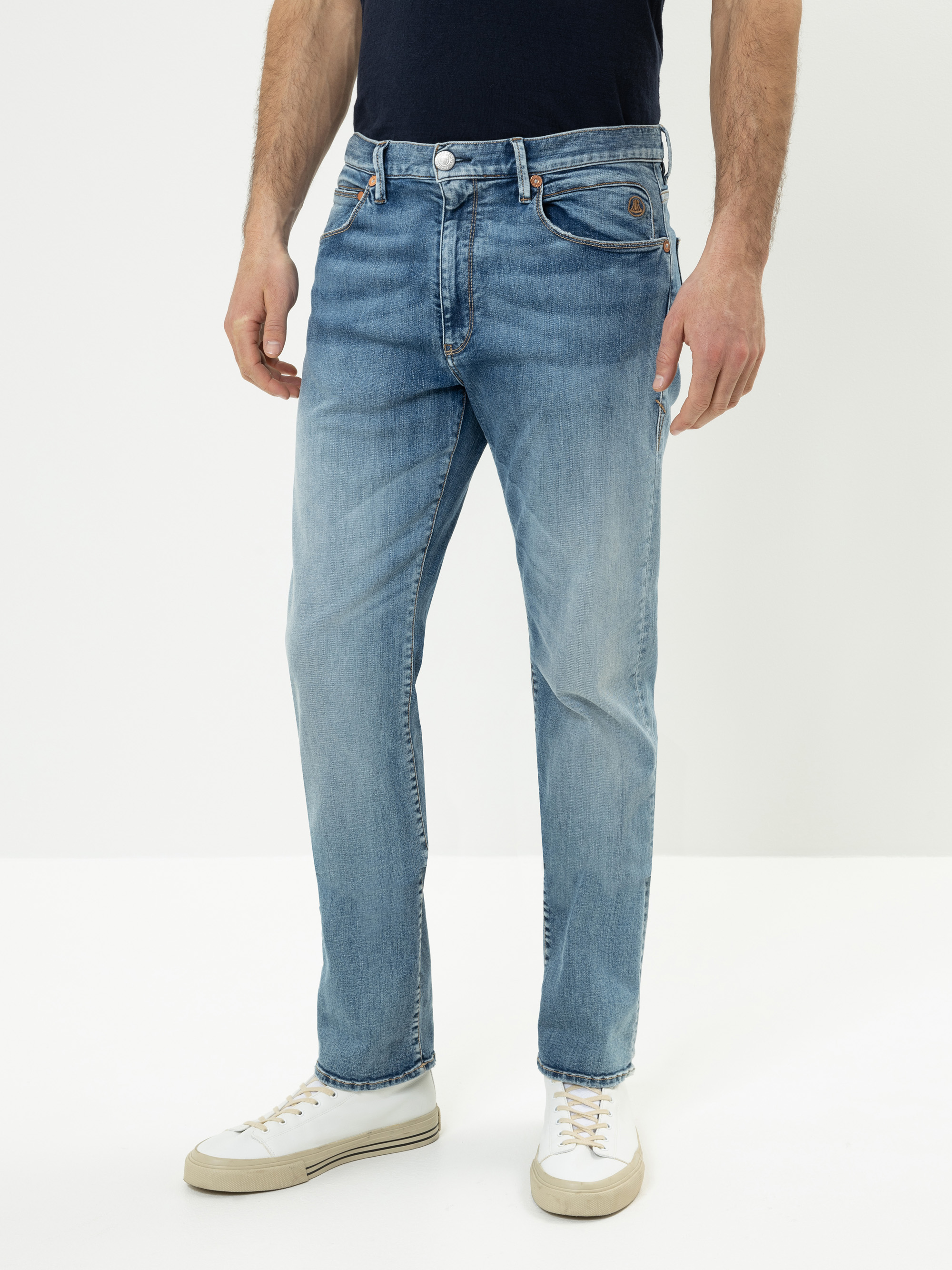 Ein Mann in hellblauen Jeans, einem dunklen T-Shirt und weißen Turnschuhen steht vor einem schlichten weißen Hintergrund. Seine Hände ruhen an den Seiten, und sein Kopf ist auf dem Bild nicht zu sehen.