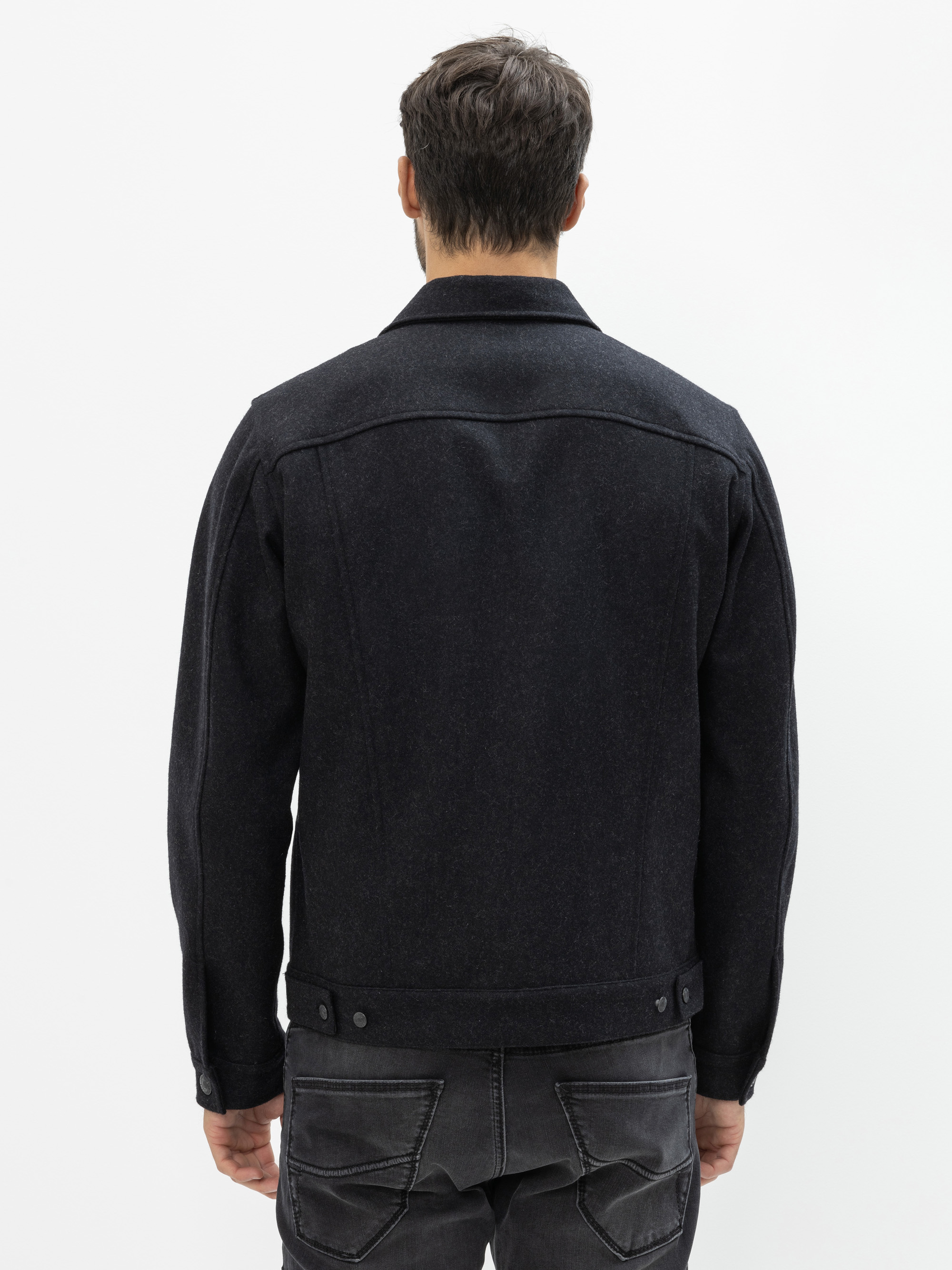 Ein Mann steht mit dem Rücken zur Kamera, trägt eine dunkle Jeansjacke und schwarze Jeans vor einem schlichten weißen Hintergrund.