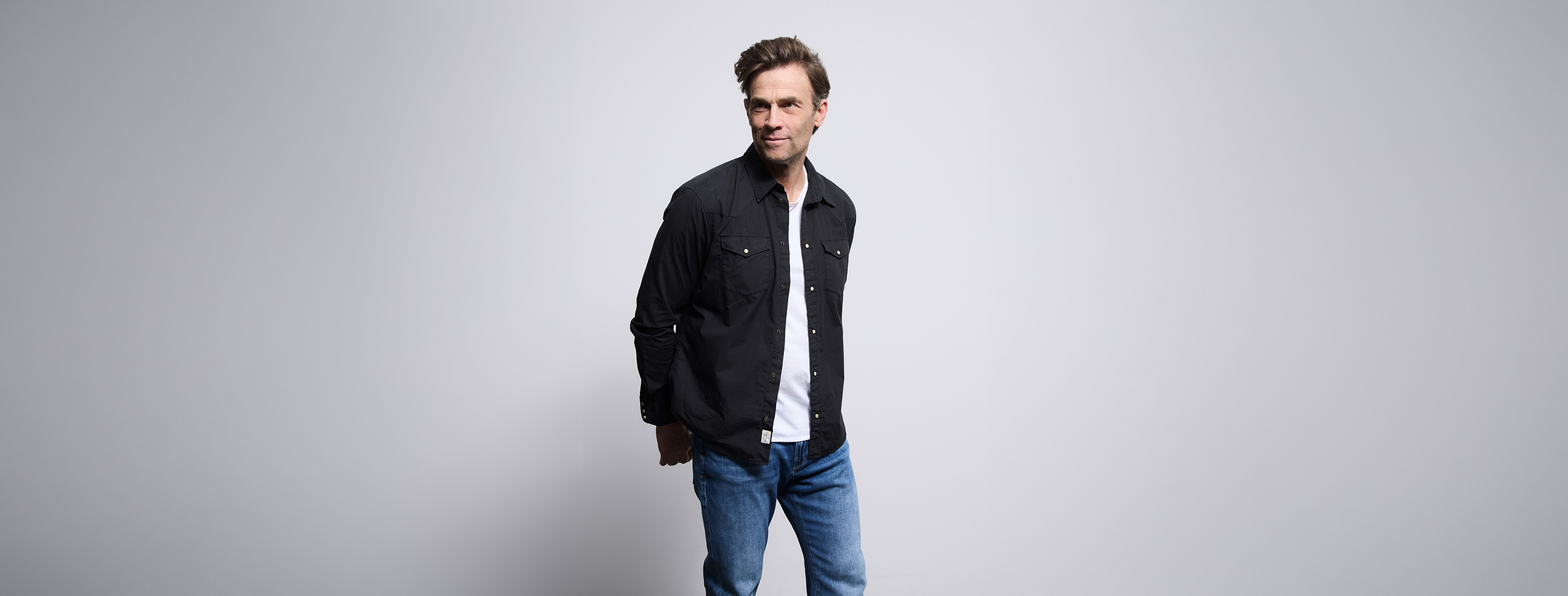 Ein Mann in einem schwarzen Button-up-Hemd über einem weißen T-Shirt und blauen Jeans steht vor einem schlichten hellgrauen Hintergrund und schaut mit einem leichten Lächeln zur Seite.