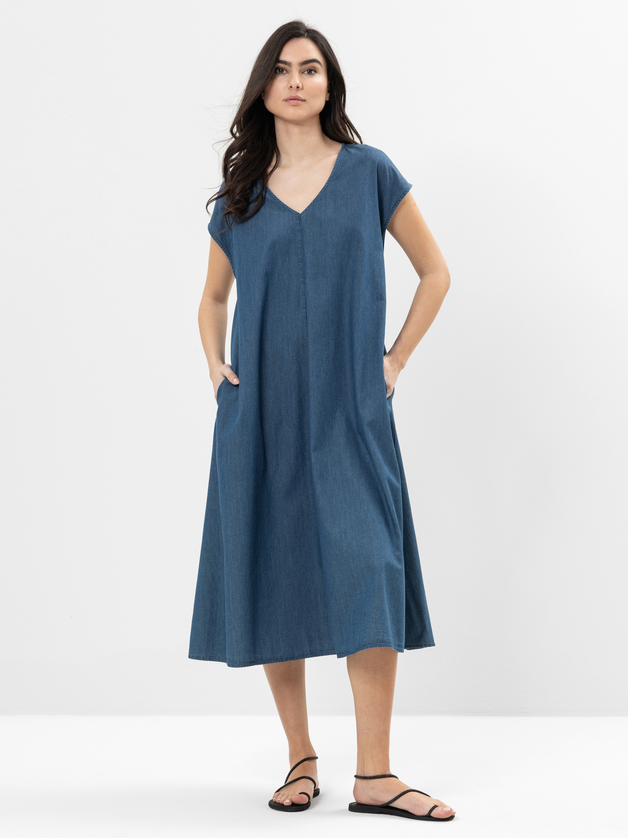 Eine Frau steht vor einem schlichten weißen Hintergrund und trägt ein locker sitzendes, kurzärmeliges, blaues Jeans-Midikleid mit V-Ausschnitt und schwarze Sandalen. Sie hat langes, gewelltes dunkles Haar und ihre Hände stecken in den Taschen des Klei