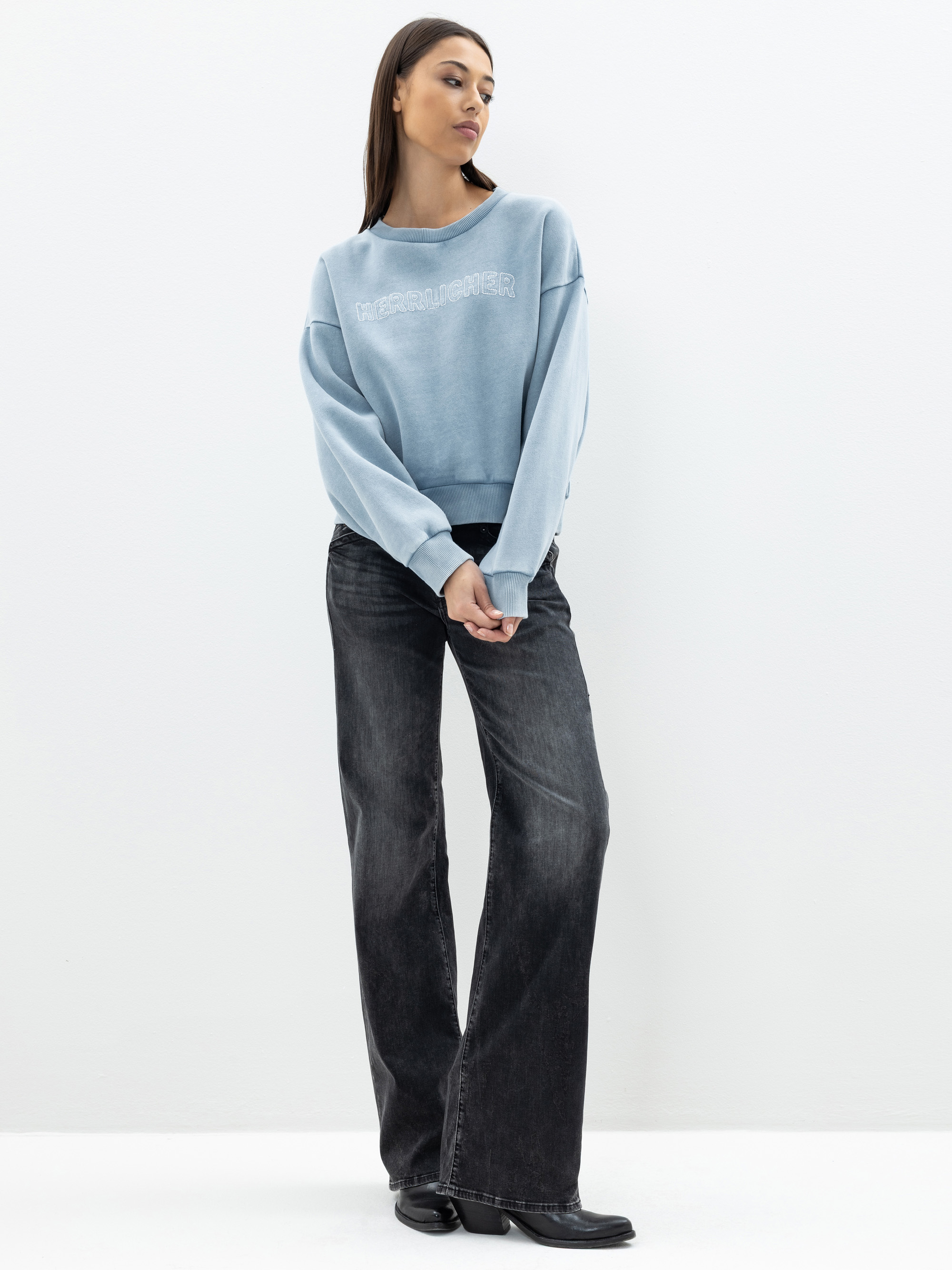 Eine Frau steht vor einem schlichten weißen Hintergrund und trägt ein hellblaues Sweatshirt mit Aufschrift, weite schwarze Jeans und schwarze Stiefel. Sie hat glattes, dunkles Haar und schaut mit vor der Brust verschränkten Händen zur Seite.