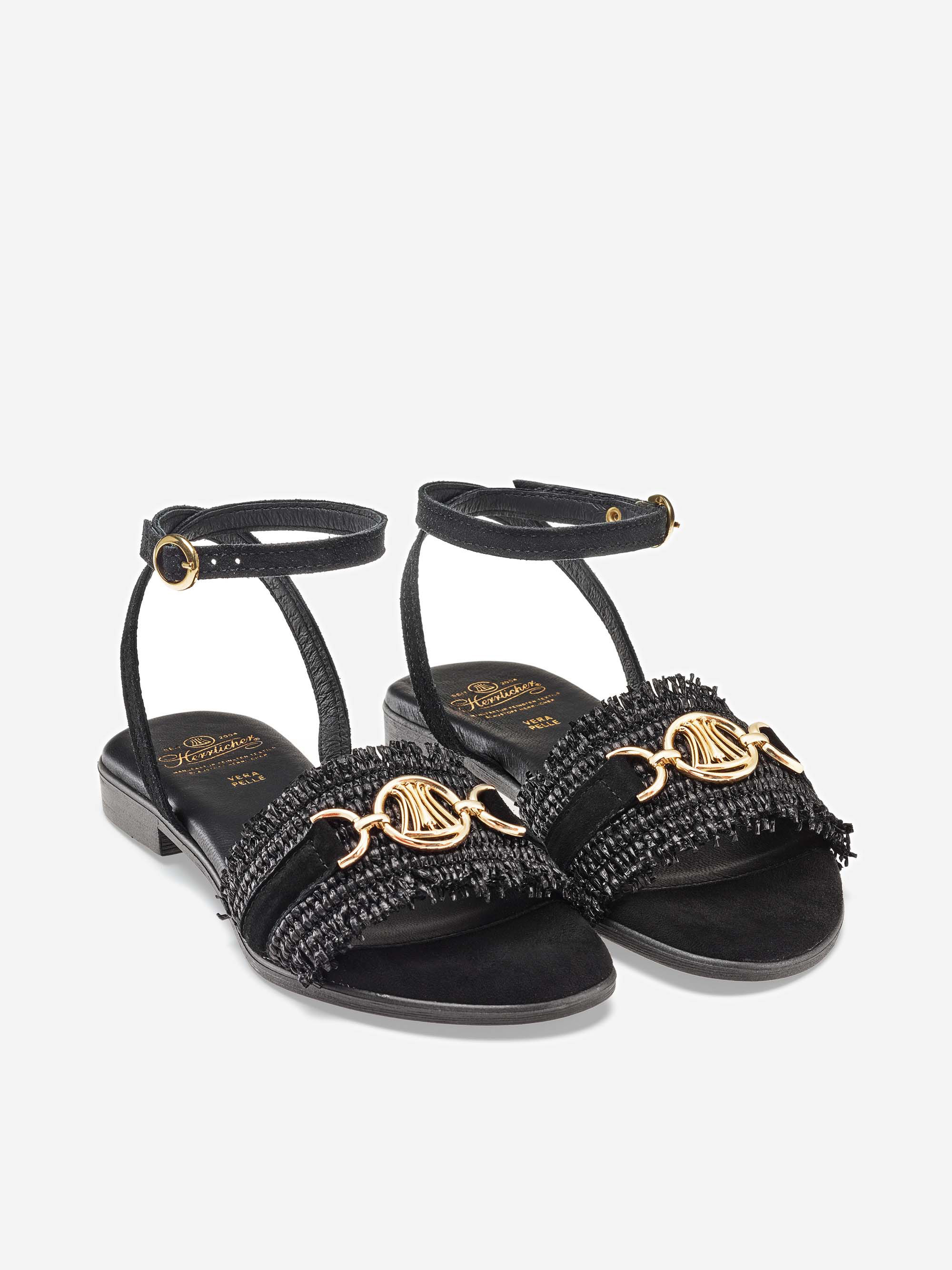 Ein Paar schwarzer, zehenoffener Sandalen mit Knöchelriemen, goldenen Schnallen, strukturierten Fransendetails und goldenen Kettenornamenten über den vorderen Riemen auf weißem Hintergrund.