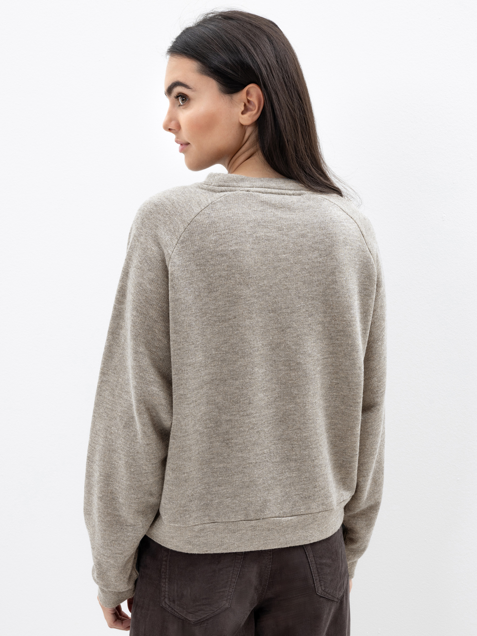 Frau mit langen dunklen Haaren, bekleidet mit einem hellgrauen Pullover und einer dunklen Hose, vor einem schlichten weißen Hintergrund stehend, den Blick abgewandt, aber den Kopf leicht zur Seite gedreht.
