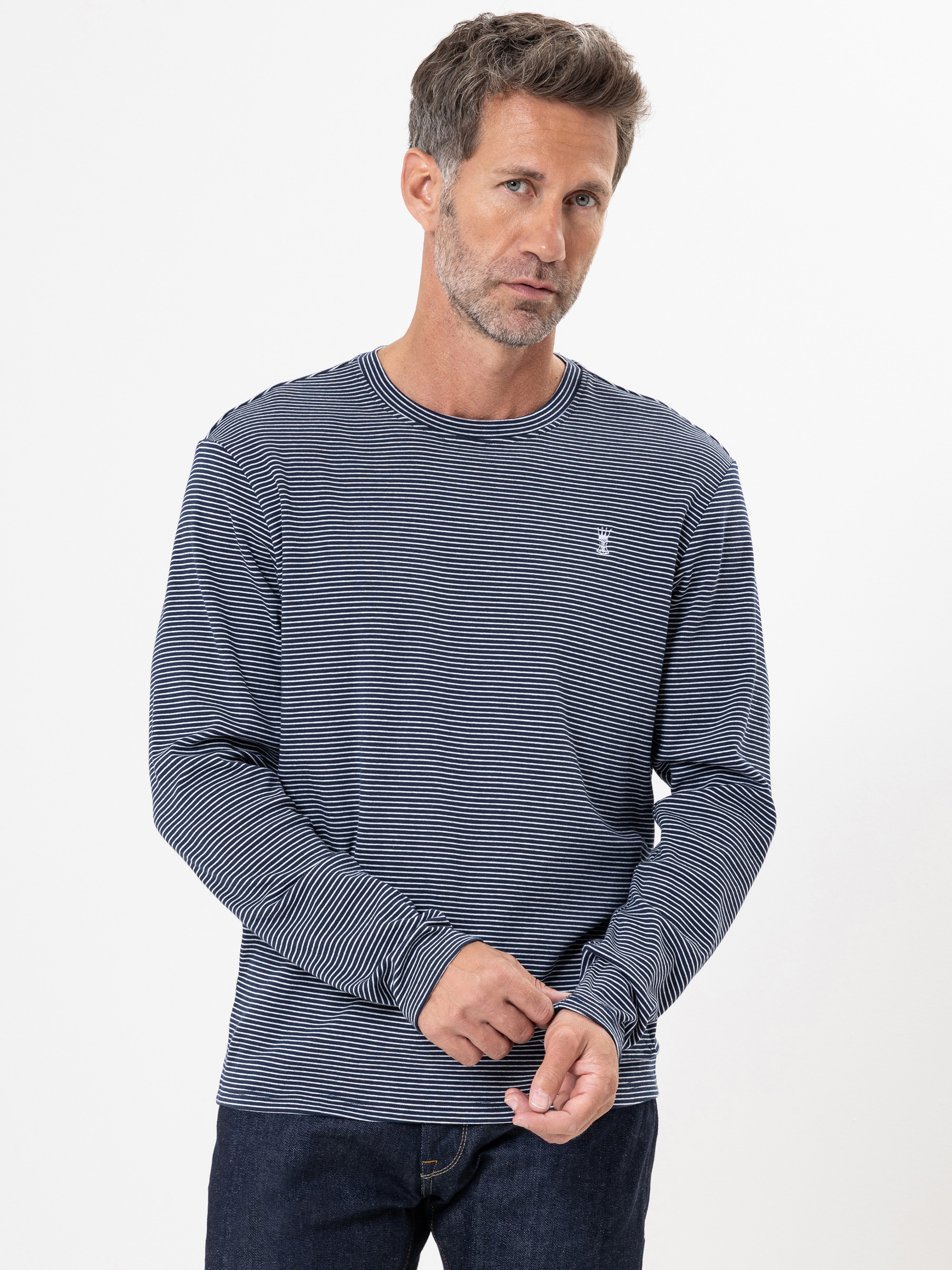 Ein Mann mit kurzem, grauem Haar und gestutztem Bart trägt ein marineblau-weiß gestreiftes, langärmeliges Hemd und dunkle Jeans und steht vor einem weißen Hintergrund. Er rückt seinen Ärmel zurecht und schaut leicht zur Seite.