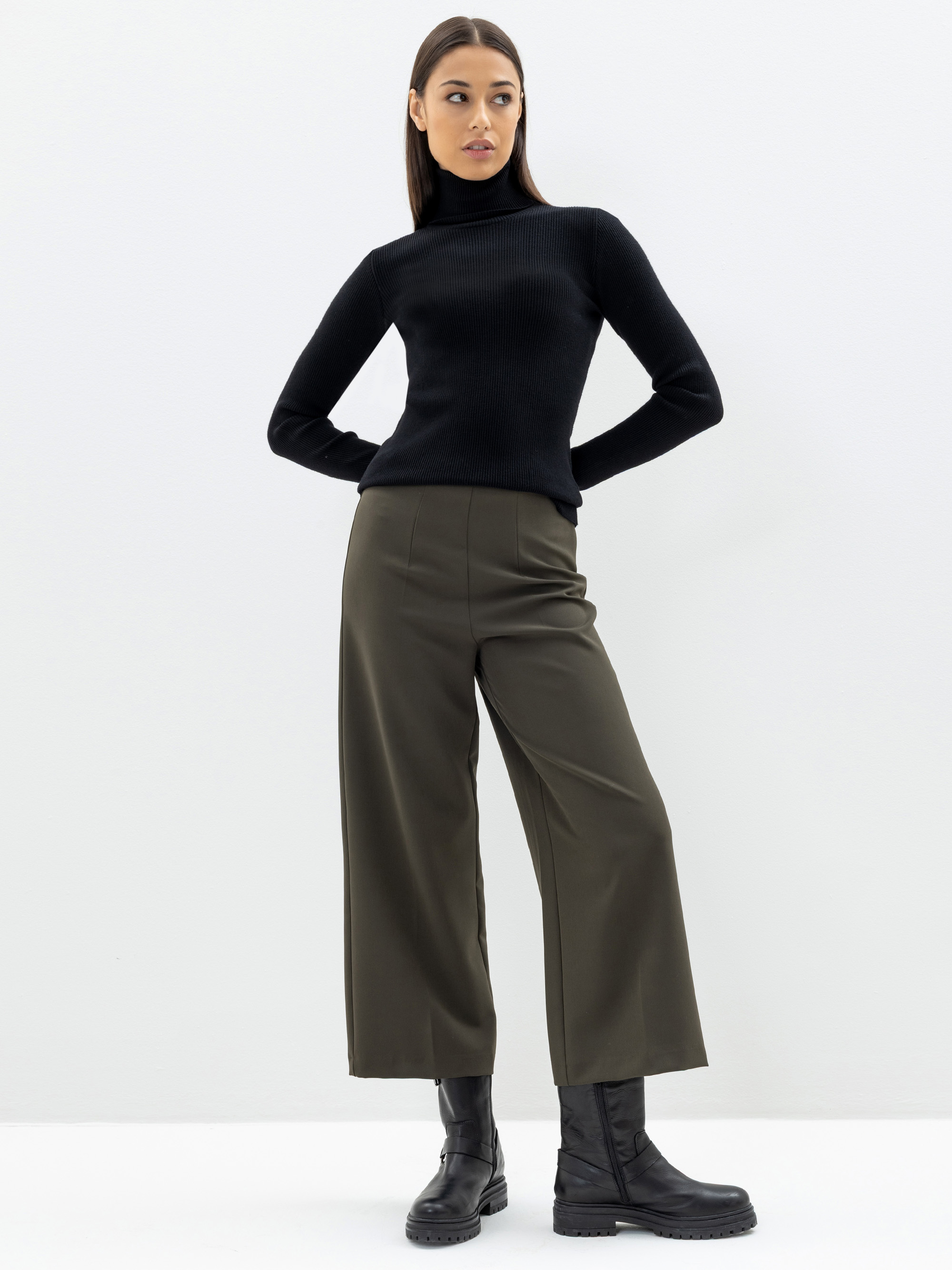 Eine Frau steht vor einem schlichten weißen Hintergrund und trägt einen schwarzen Rollkragenpullover, eine olivgrüne Hose mit weitem Bein und schwarze Stiefel. Ihre Hände ruhen auf den Hüften und sie blickt leicht zur Seite.
