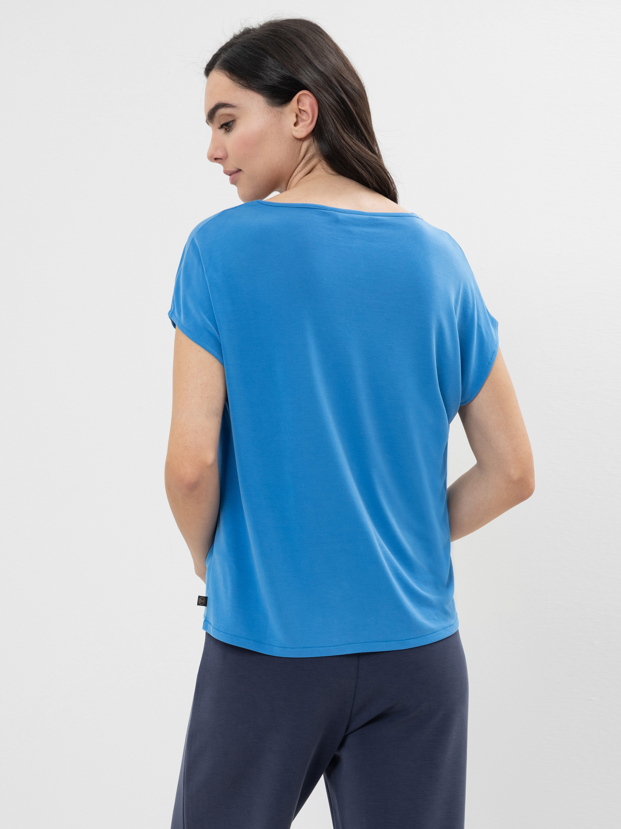 Eine Frau mit langen dunklen Haaren steht mit dem Rücken zur Kamera, trägt ein locker sitzendes blaues T-Shirt und eine marineblaue Hose vor einem schlichten, hellen Hintergrund.