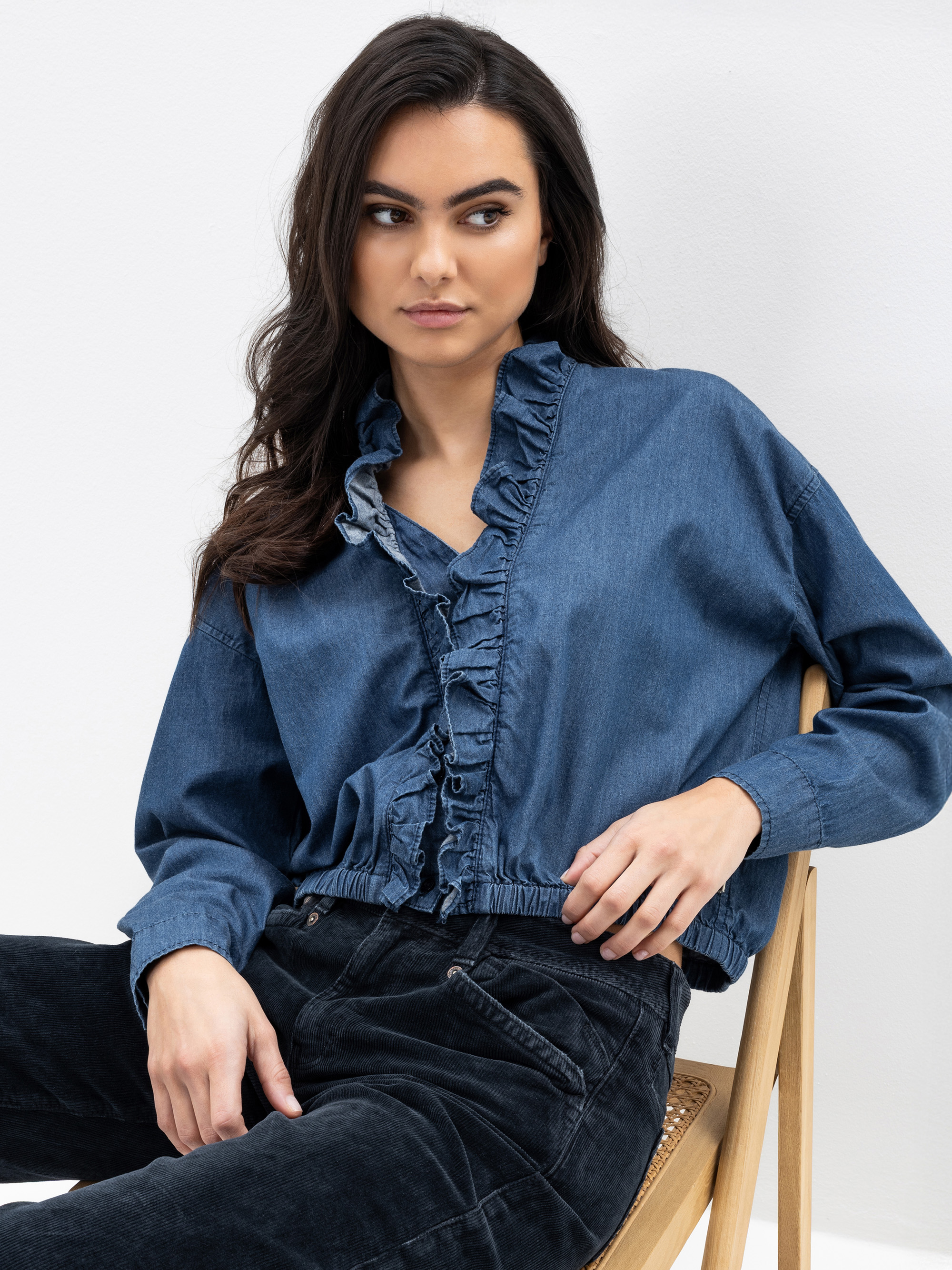 Eine Frau mit langen dunklen Haaren sitzt auf einem Holzstuhl und trägt eine blaue Jeansbluse mit Rüschenbesatz und schwarze Jeans. Sie blickt mit neutraler Miene vor einem schlichten weißen Hintergrund zur Seite.
