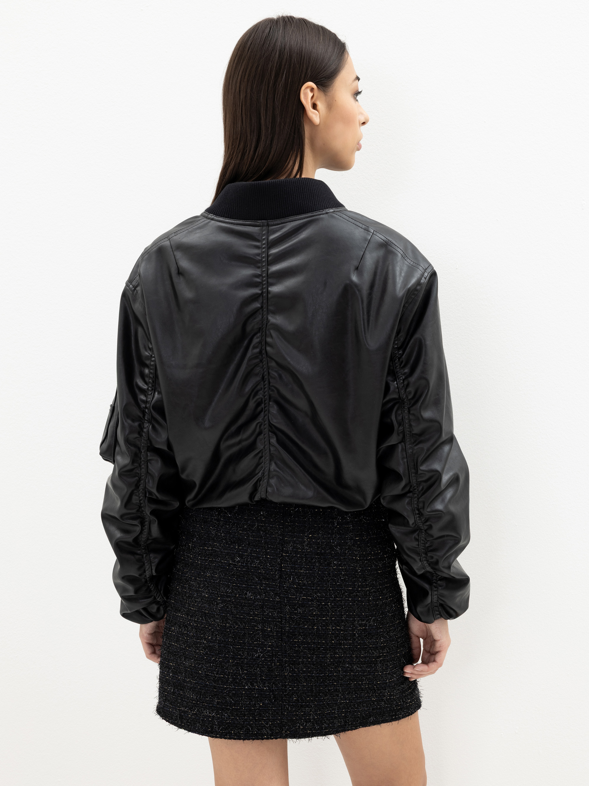 Eine Frau mit glattem dunklem Haar, von hinten gesehen, trägt eine schwarze Lederjacke mit gerüschten Ärmeln und einen schwarzen strukturierten Rock. Sie steht vor einem schlichten weißen Hintergrund.
