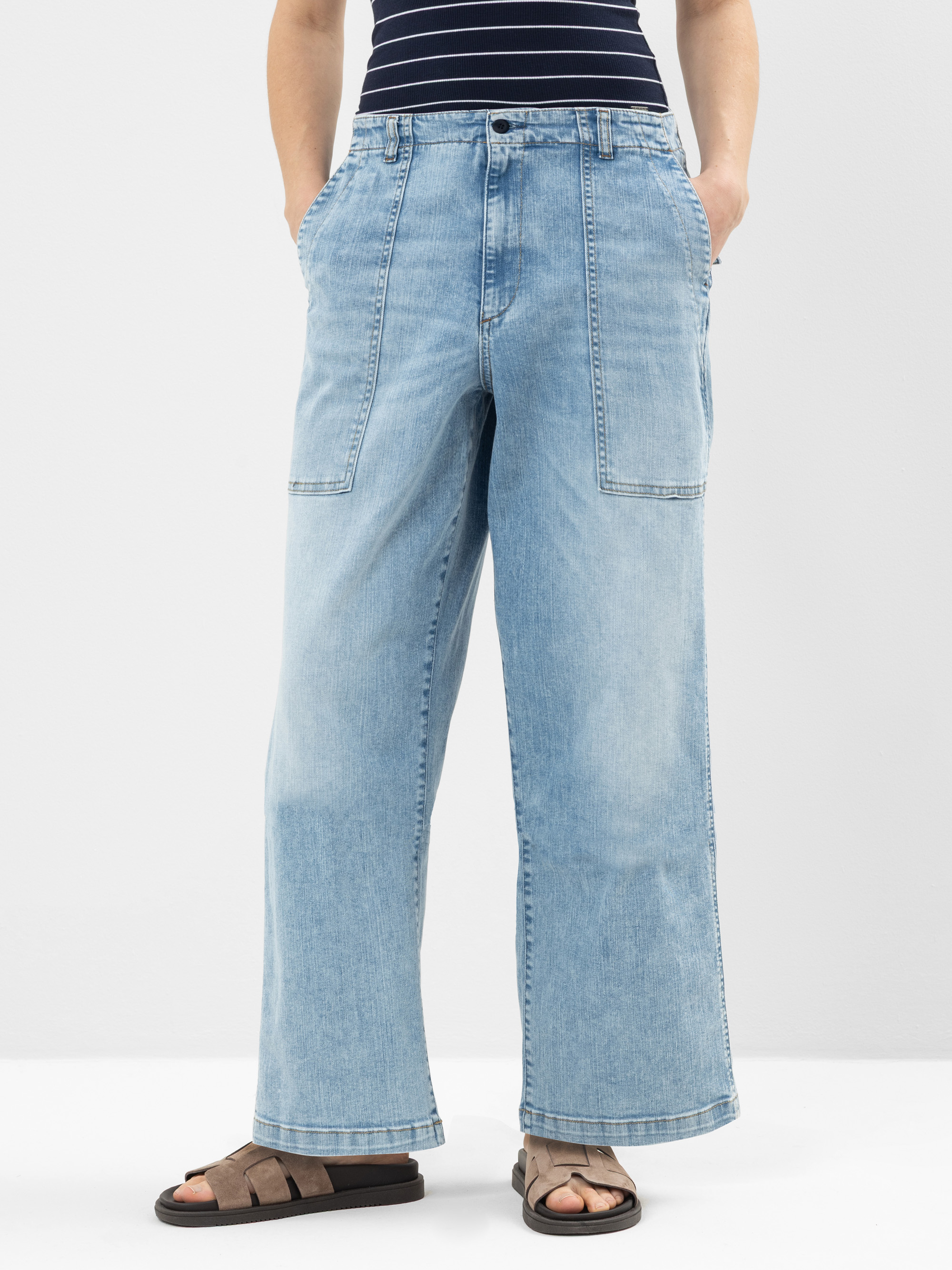 Person, die hellblaue Jeans mit weitem Bein und großen Vordertaschen, ein marineblau-weiß gestreiftes Oberteil und braune Sandalen trägt und vor einem schlichten hellen Hintergrund steht. Die Hände stecken in den Taschen der Jeans.