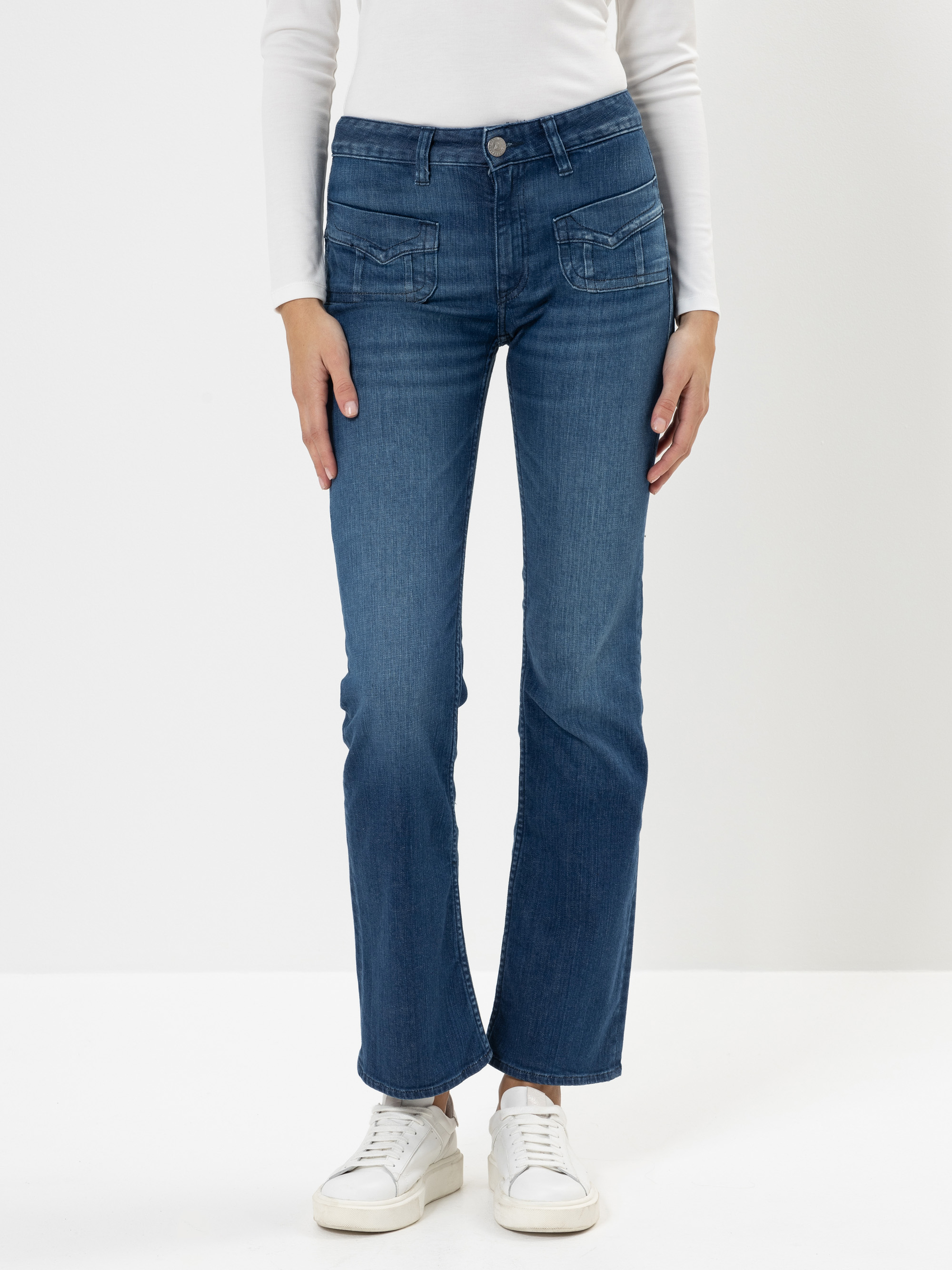 Eine Person, die blaue Jeans, ein langärmeliges weißes Oberteil und weiße Turnschuhe trägt, steht vor einem schlichten weißen Hintergrund. Nur der Körper von den Schultern abwärts ist sichtbar.