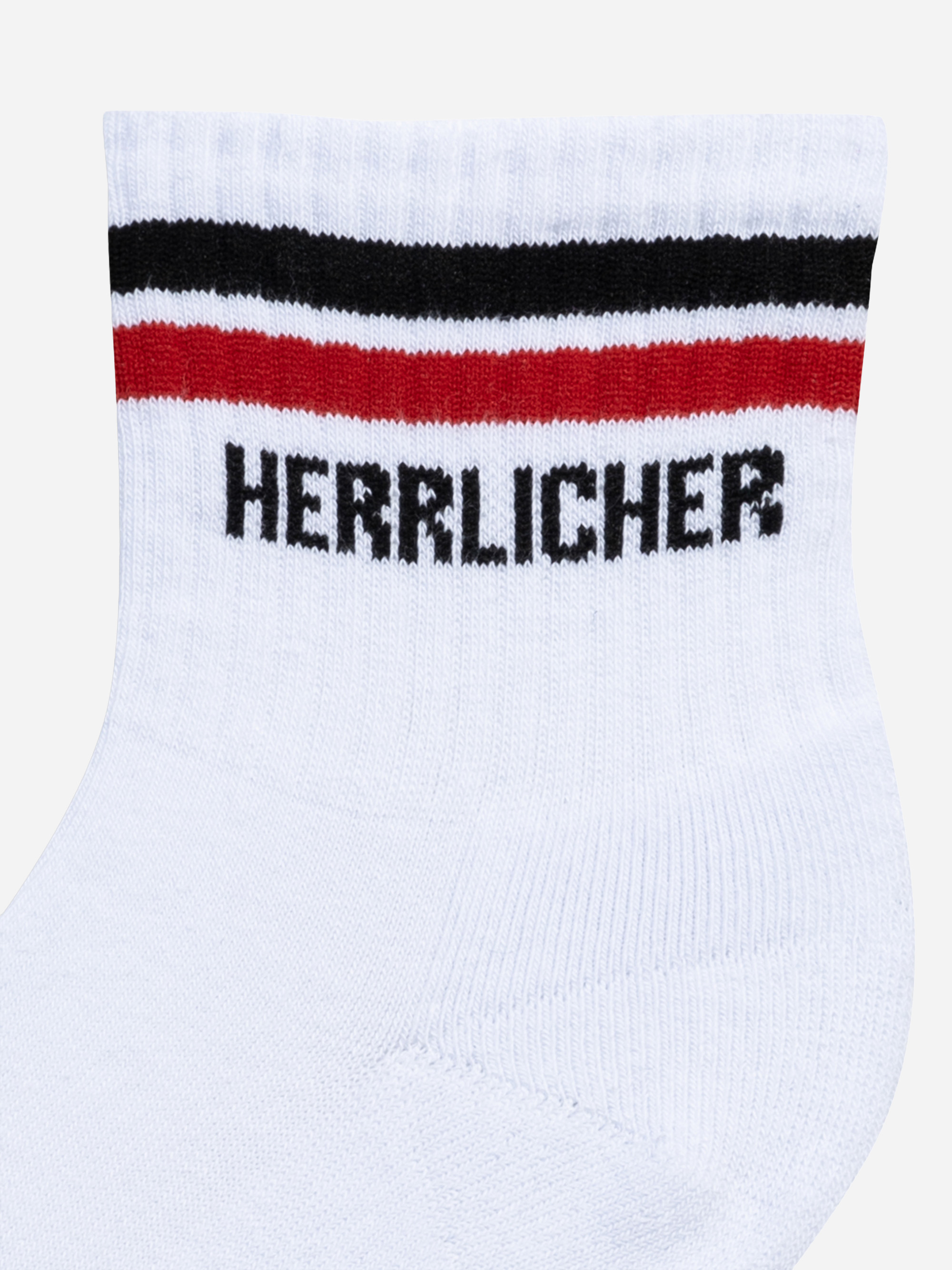 Nahaufnahme einer weißen Socke mit zwei horizontalen Streifen, einem schwarzen und einem roten, in der Nähe des Bündchens, und dem Wort HERRLICHER in schwarzen, fetten Buchstaben unter den Streifen.