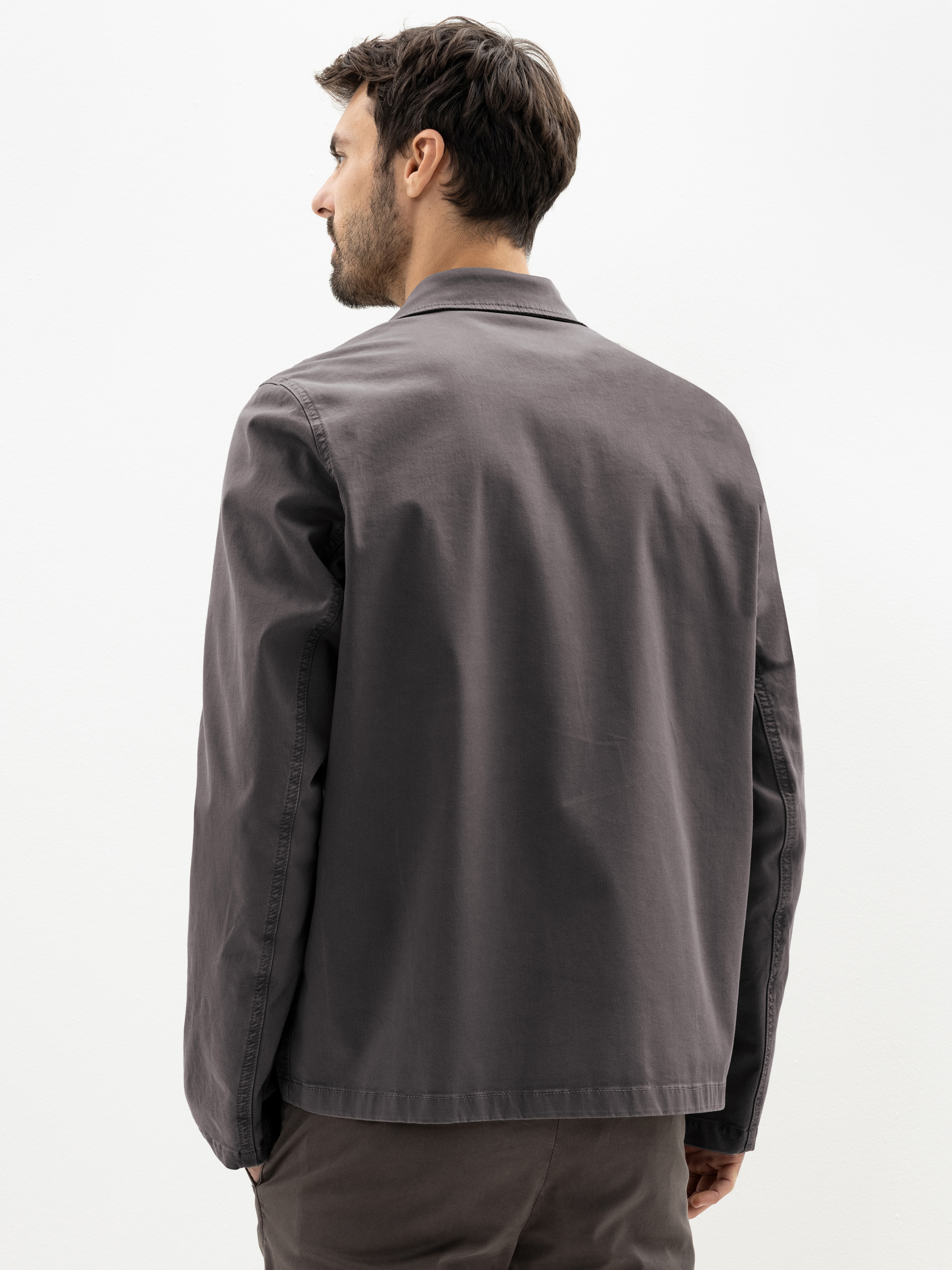 Ein Mann mit kurzen dunklen Haaren steht, von der Kamera abgewandt, in einer grauen Jacke mit Kragen und einer passenden grauen Hose vor einem schlichten weißen Hintergrund.