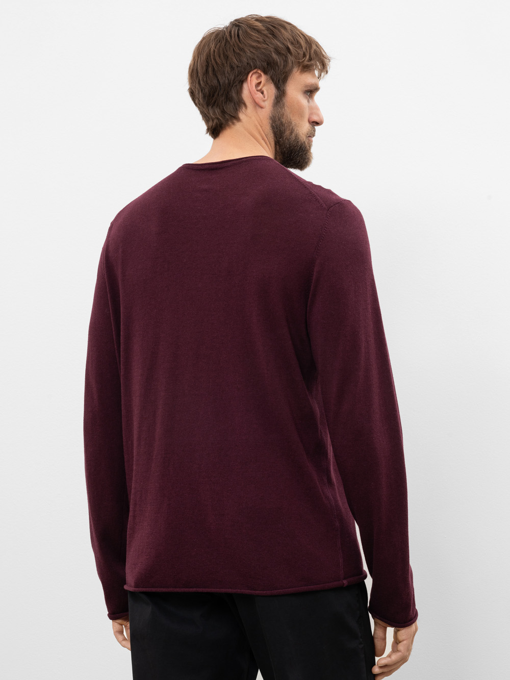 Ein Mann mit kurzen braunen Haaren und einem Bart steht, von der Kamera abgewandt, in einem langärmeligen, weinroten Hemd und einer schwarzen Hose vor einem schlichten weißen Hintergrund.