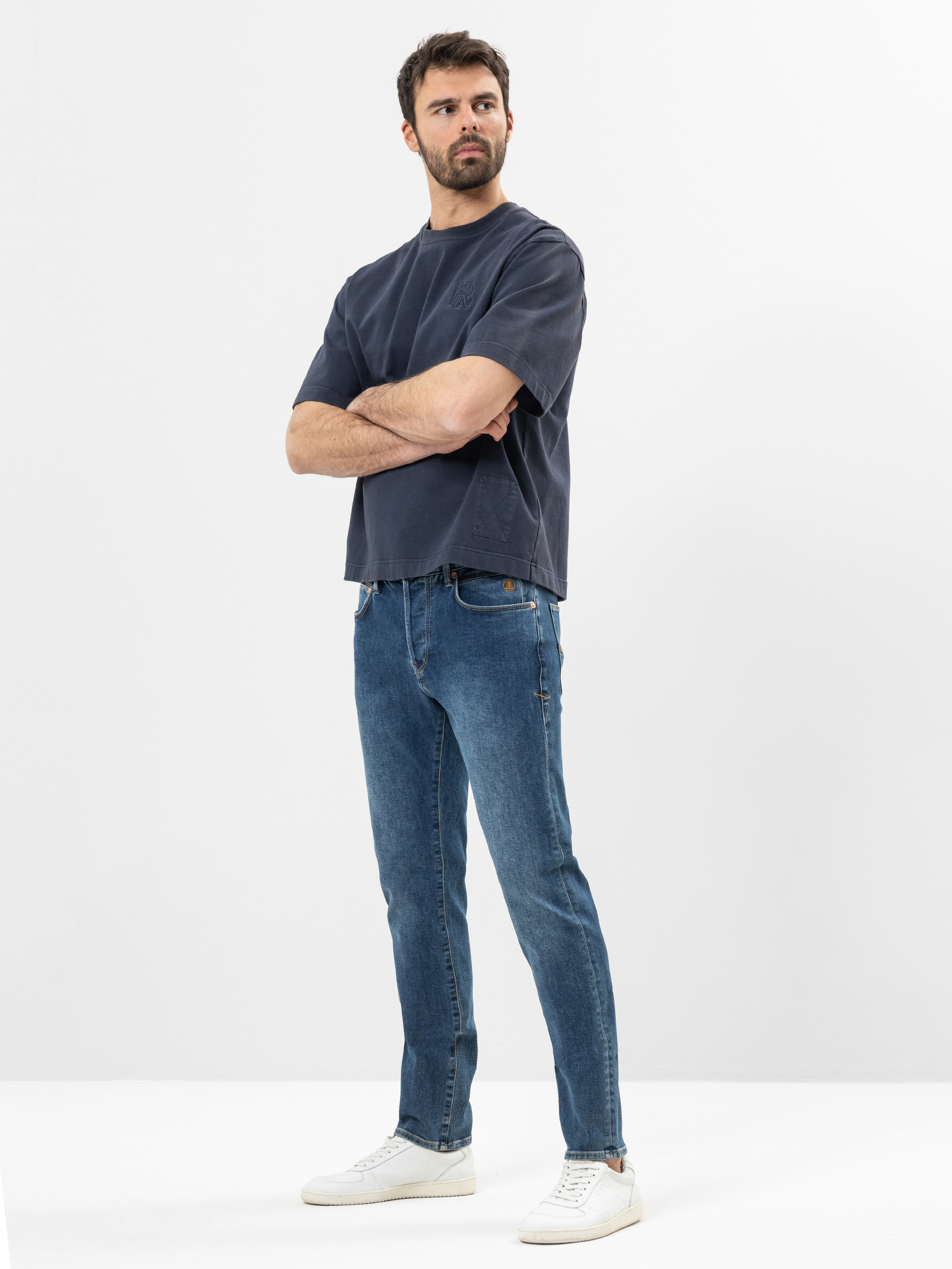 Ein Mann mit kurzen dunklen Haaren und einem Bart steht mit verschränkten Armen, einem lockeren dunklen T-Shirt, blauen Jeans und weißen Turnschuhen vor einem schlichten weißen Hintergrund.