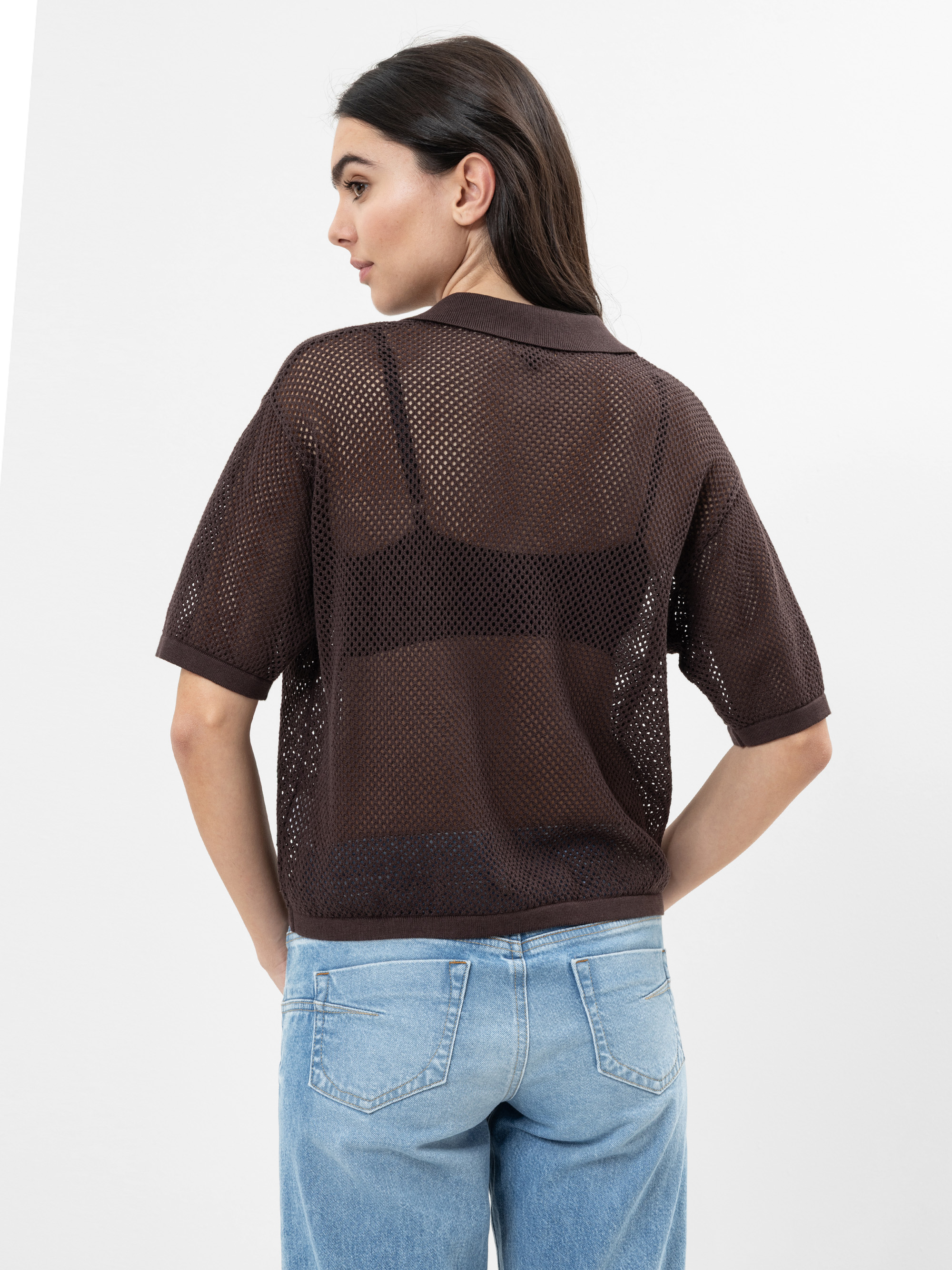Eine Frau posiert mit dem Rücken zur Kamera. Sie trägt ein durchsichtiges, kurzärmeliges, braunes Netzhemd über einem schwarzen BH und hellblaue Jeans. Ihr dunkles Haar ist glatt und offen, und sie steht vor einem schlichten weißen Hintergrund.