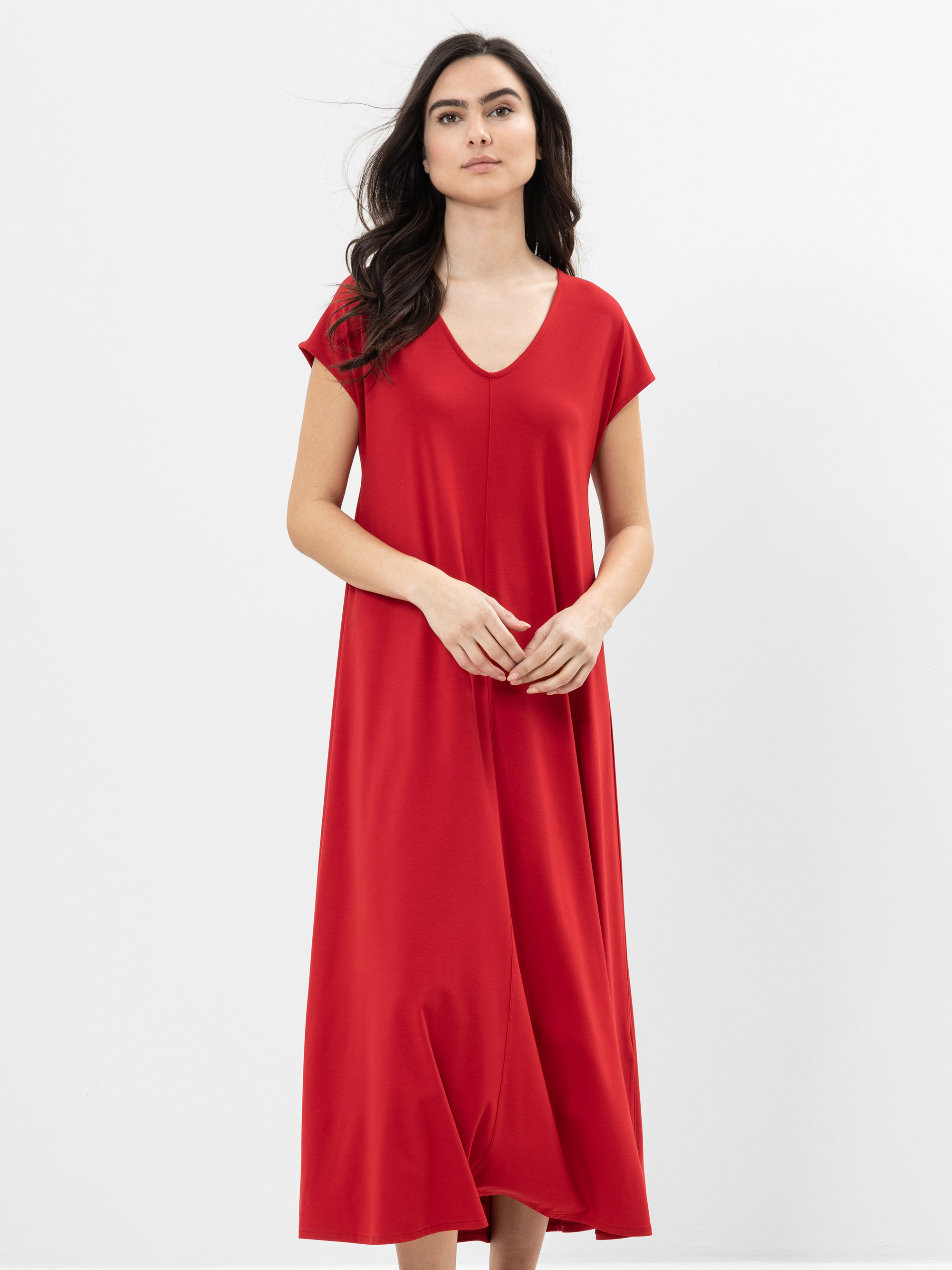 Eine Frau mit langen dunklen Haaren, die ein locker sitzendes, kurzärmeliges, rotes Kleid mit V-Ausschnitt trägt, steht vor einem schlichten weißen Hintergrund.
