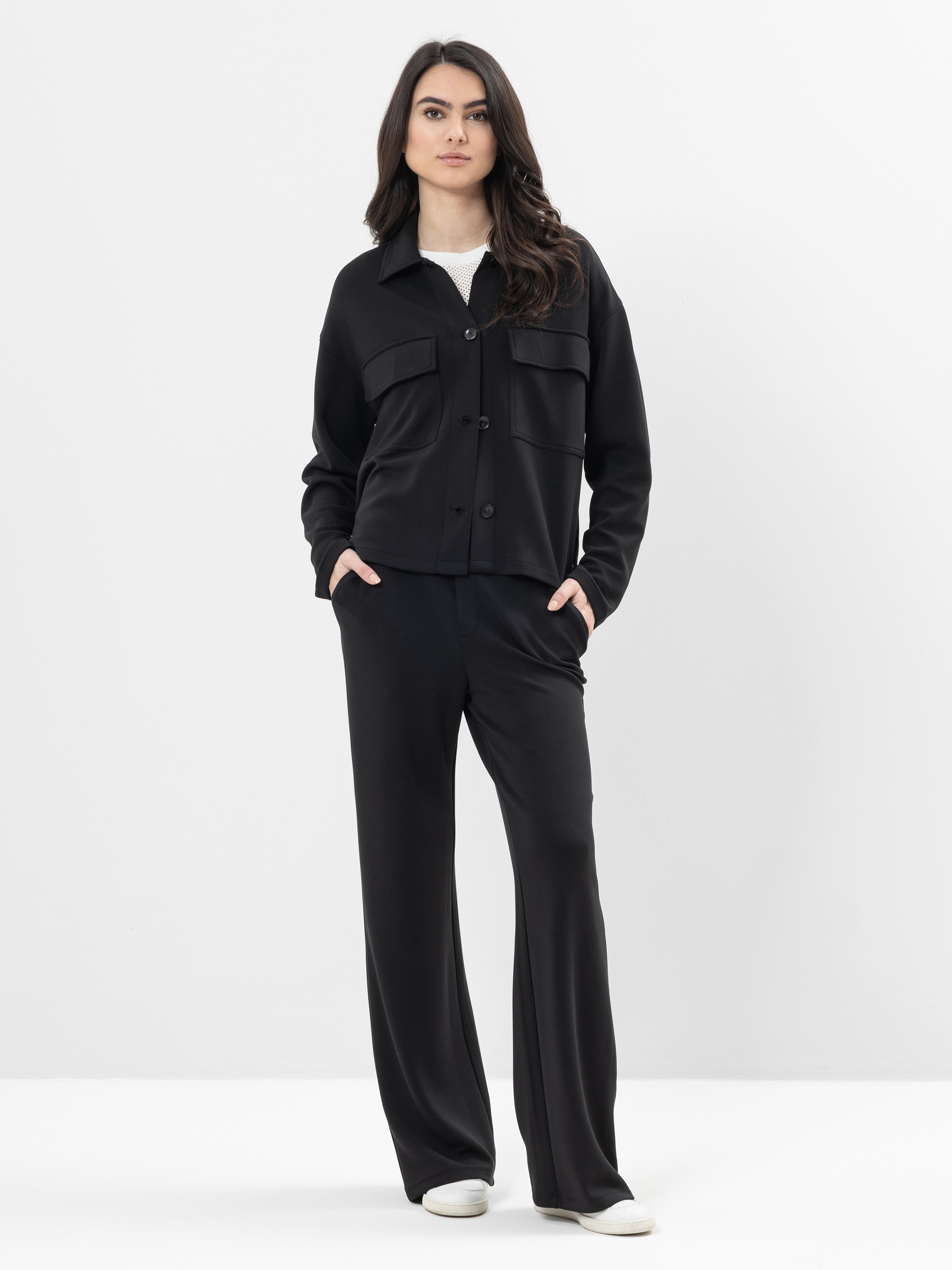 Eine Frau steht vor einem schlichten weißen Hintergrund und trägt eine schwarze Button-up-Jacke, eine dazu passende schwarze Hose mit weitem Bein, ein weißes Top und weiße Turnschuhe. Sie hat die Hände in den Taschen und blickt selbstbewusst in die K