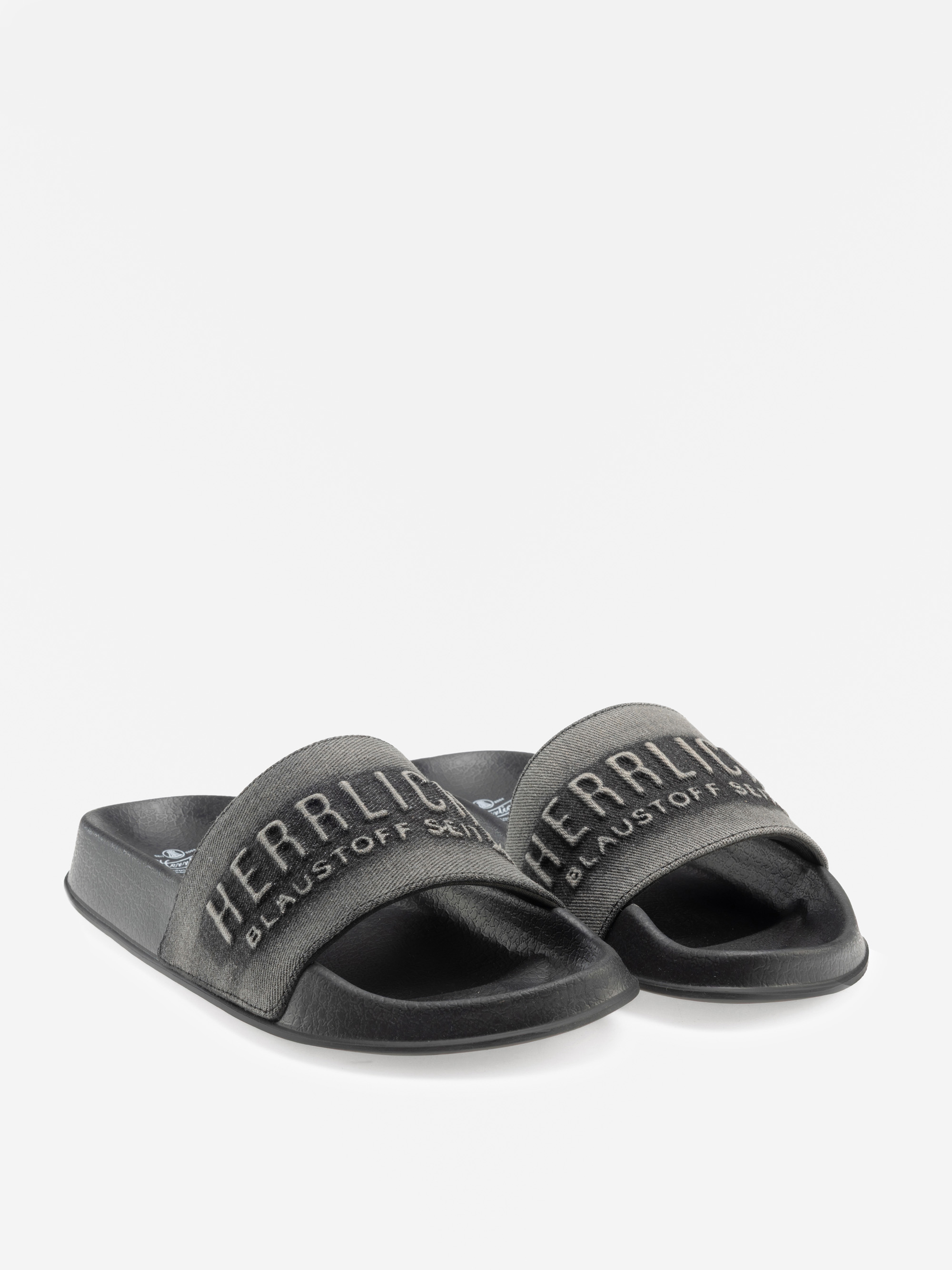 Ein Paar schwarze Gleitsandalen mit breiten Riemen. Auf den Riemen sind in weißer Schrift die Worte HERRLICH und BLAUSTOFF SCH zu lesen. Die Sandalen sind auf einem schlichten weißen Hintergrund abgebildet.