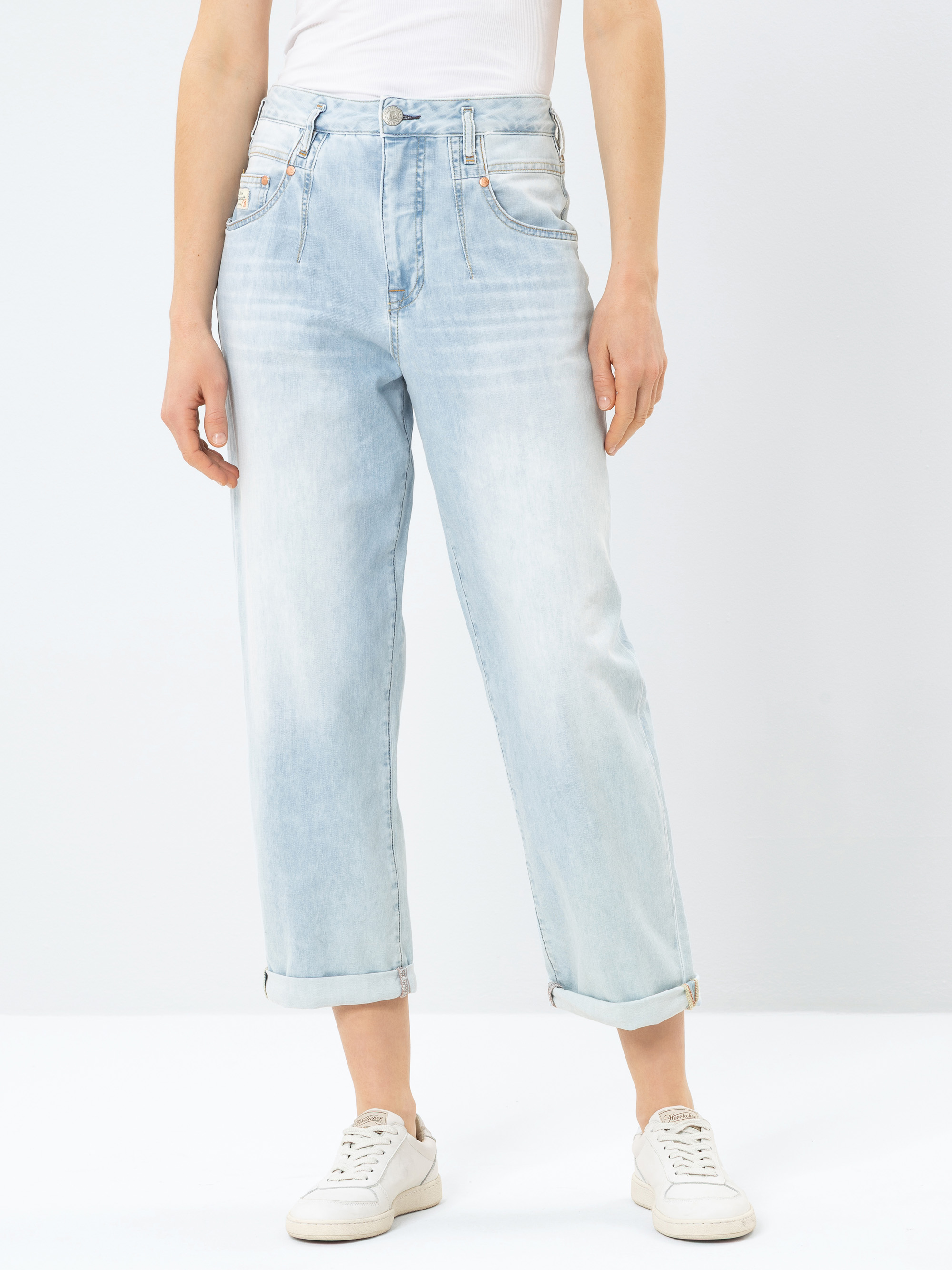 Person, die eine hellblaue Jeans mit hoher Taille und geradem Bein mit Bündchen an den Knöcheln, ein weißes, in die Hose gestecktes Oberteil und weiße Turnschuhe trägt und vor einem schlichten hellen Hintergrund steht.