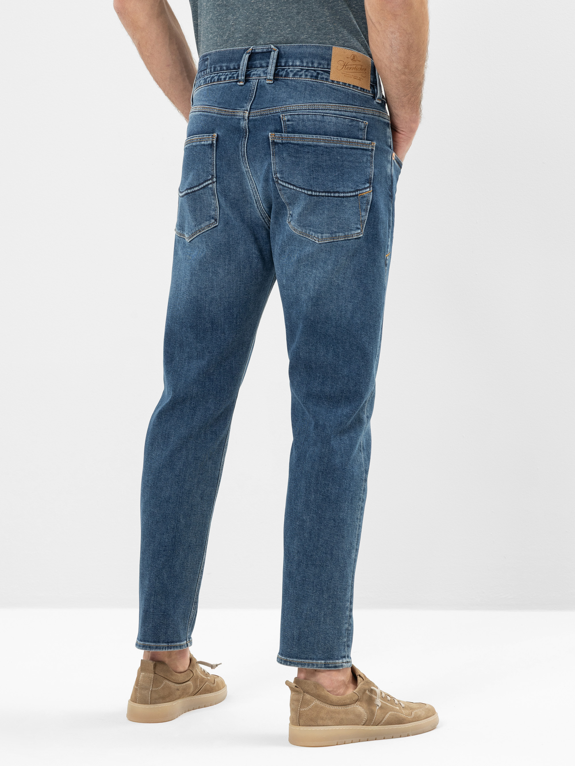 Eine Person, die eine blaue Jeans, ein graues T-Shirt und hellbraune Turnschuhe trägt, steht mit den Händen in den Taschen, von der Kamera abgewandt, vor einem schlichten weißen Hintergrund.