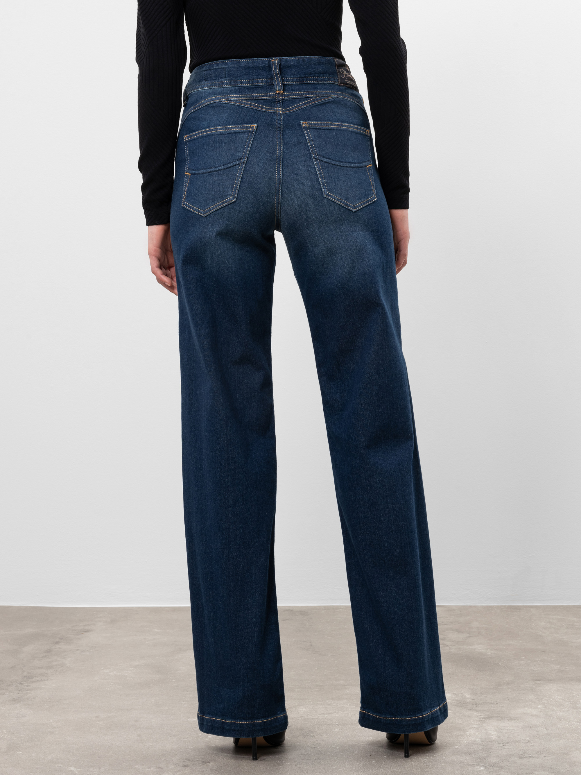 Eine Person, die eine hochgeschnittene, weit geschnittene Jeans und ein langärmeliges schwarzes Oberteil trägt, steht auf einem Betonboden, der von hinten gegen eine schlichte weiße Wand zeigt.