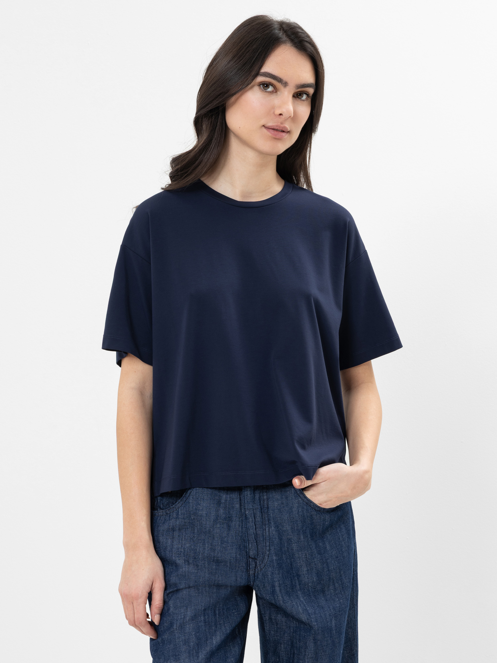 Eine Frau mit langen dunklen Haaren trägt ein marineblaues T-Shirt und blaue Jeans. Sie steht vor einem schlichten weißen Hintergrund, hat eine Hand in der Tasche und einen ruhigen Gesichtsausdruck.