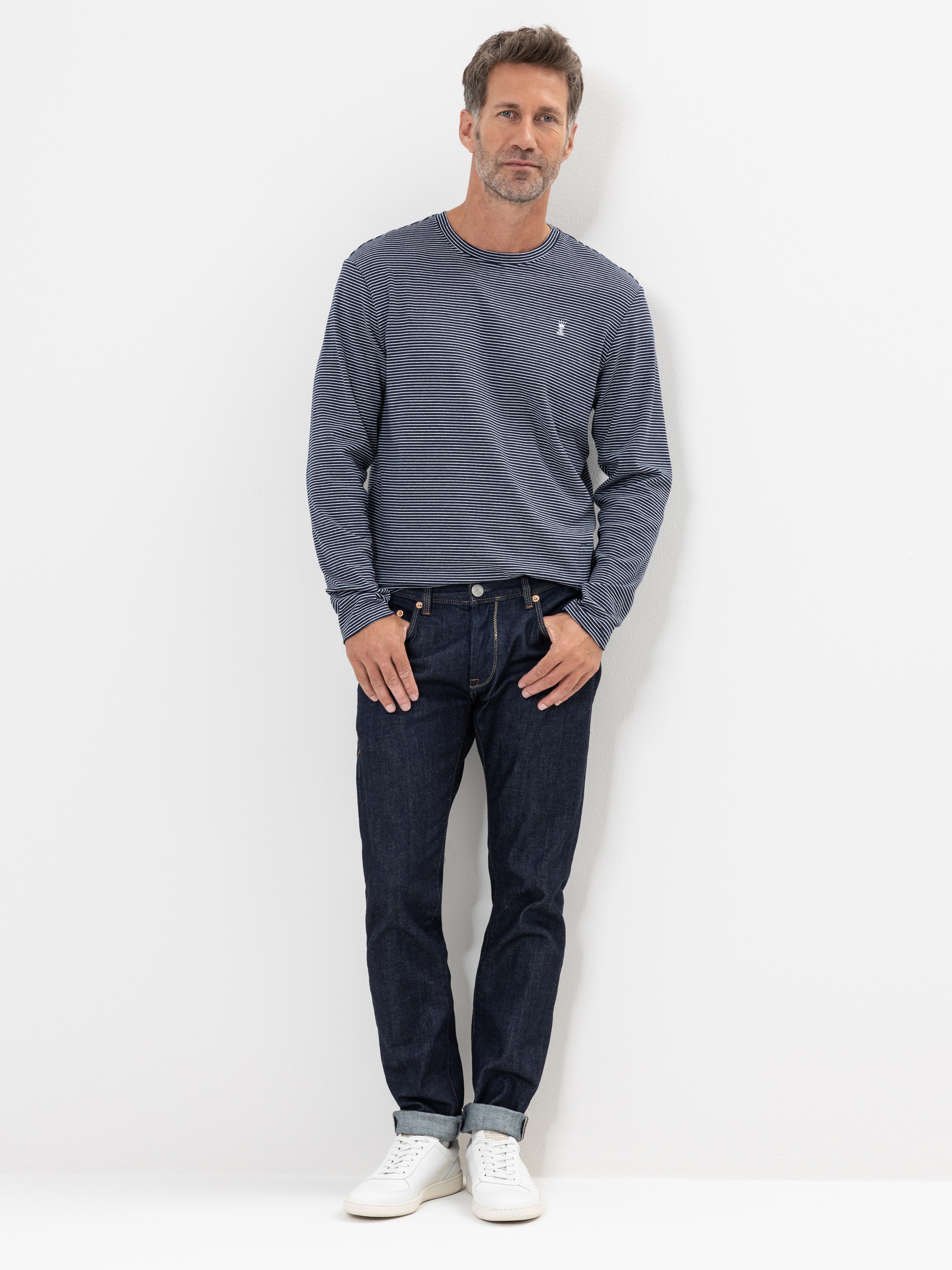 Ein Mann steht vor einem weißen Hintergrund und trägt ein blaues langärmeliges Hemd, dunkle Jeans und weiße Turnschuhe. Er hat kurzes braunes Haar und einen gestutzten Bart. Er hat die linke Hand in der Tasche und einen entspannten Gesichtsausdruck.