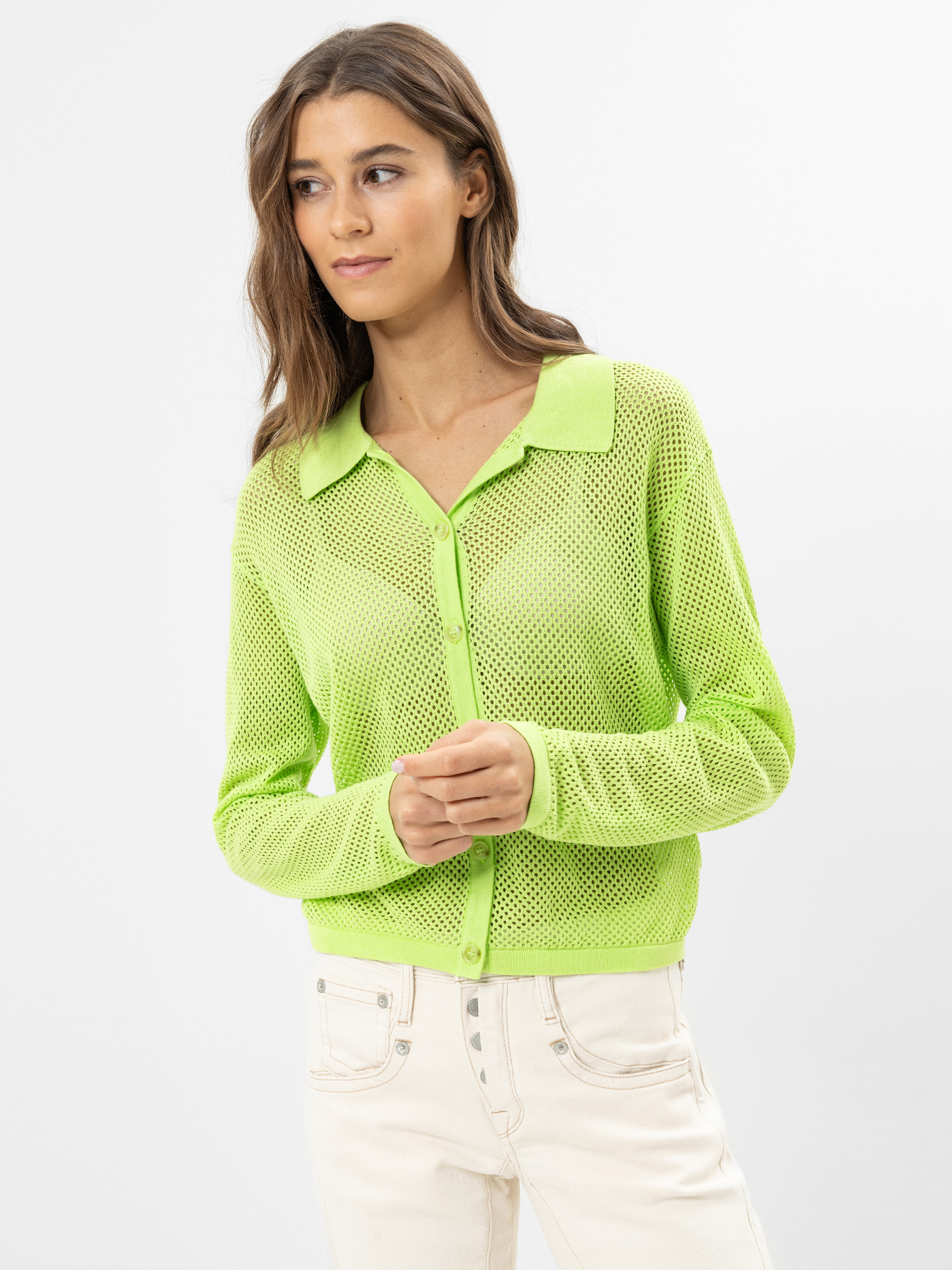 Eine Frau mit langen braunen Haaren trägt eine hellgrüne, geknüpfte Strickjacke über einem blassgrünen Oberteil und hellbeigefarbene Jeans vor einem schlichten weißen Hintergrund.