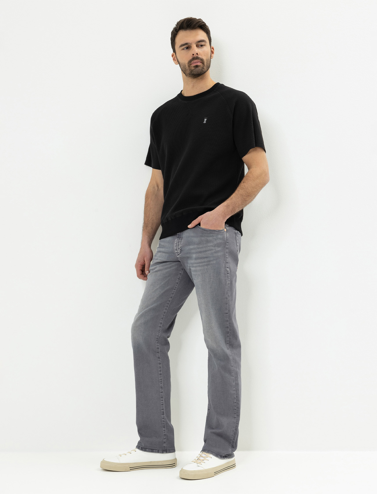 Ein Mann steht vor einem weißen Hintergrund und trägt ein schwarzes kurzärmeliges T-Shirt, hellgraue Jeans und weiße Turnschuhe. Er hat eine Hand in der Tasche und einen neutralen Gesichtsausdruck.