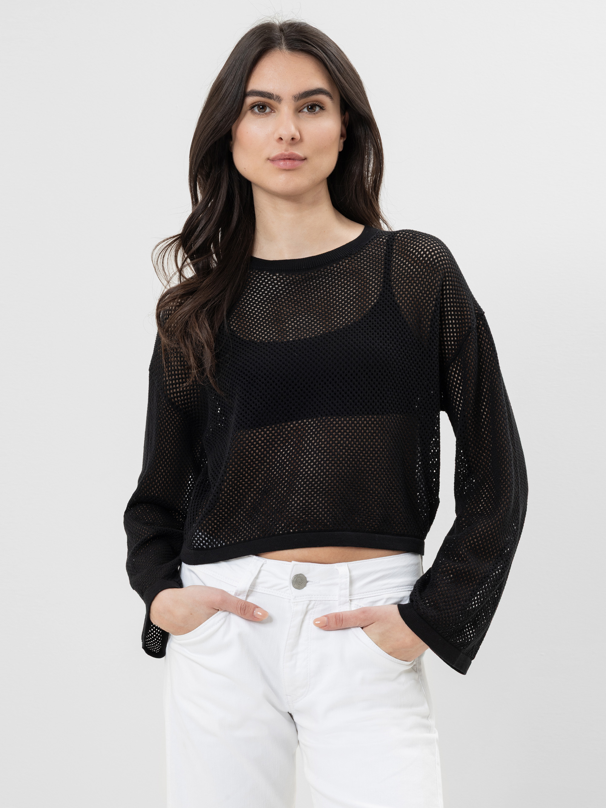 Eine Frau mit langen dunklen Haaren trägt ein schwarzes langärmeliges Netzoberteil über einem schwarzen BH und eine weiße Hose mit hoher Taille. Sie steht mit den Händen in den Taschen vor einem schlichten hellen Hintergrund.