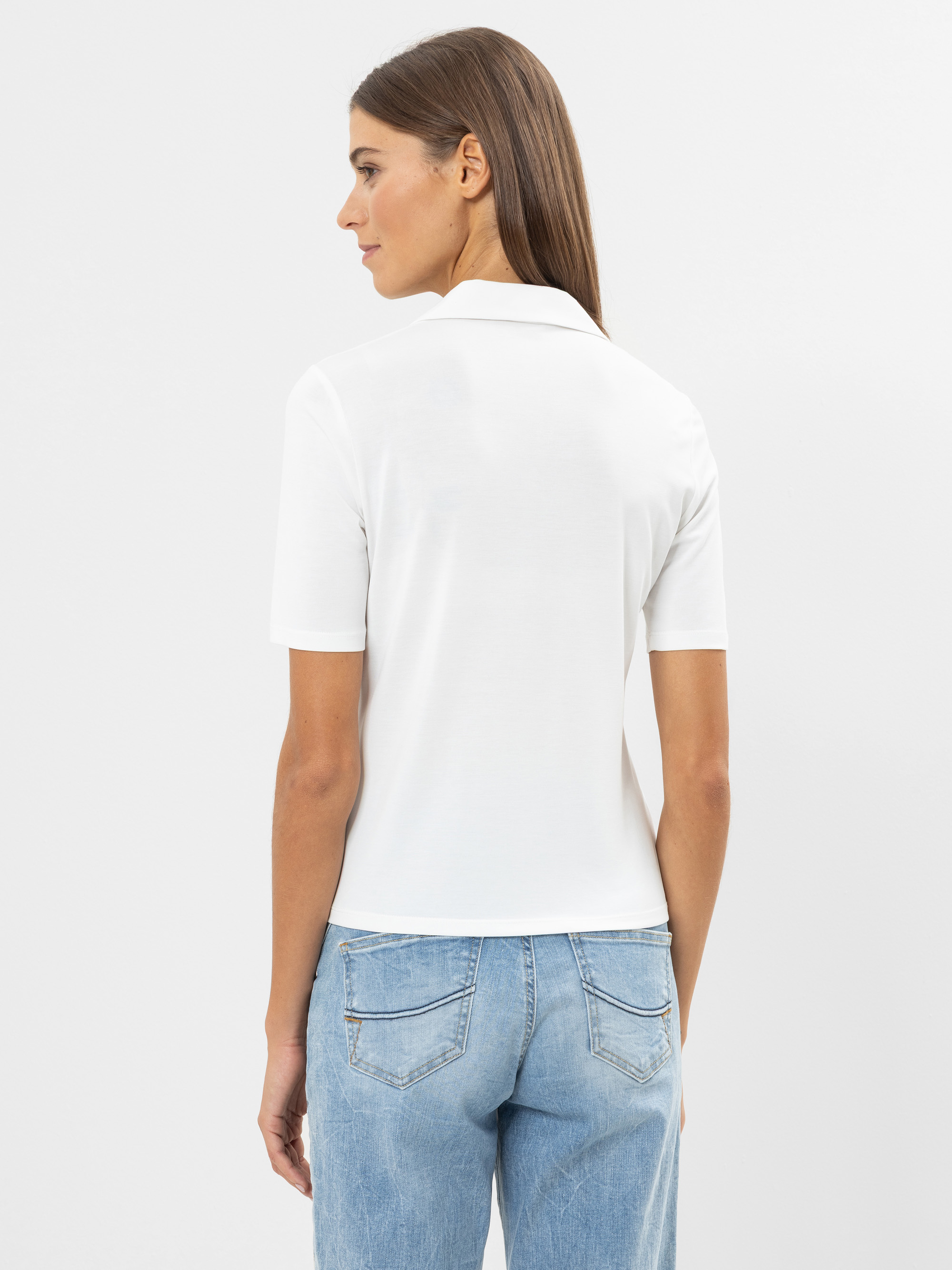 Eine Frau mit glattem braunem Haar wird von hinten gezeigt. Sie trägt ein weißes kurzärmeliges Hemd mit Kragen und hellblaue Jeans vor einem schlichten weißen Hintergrund.