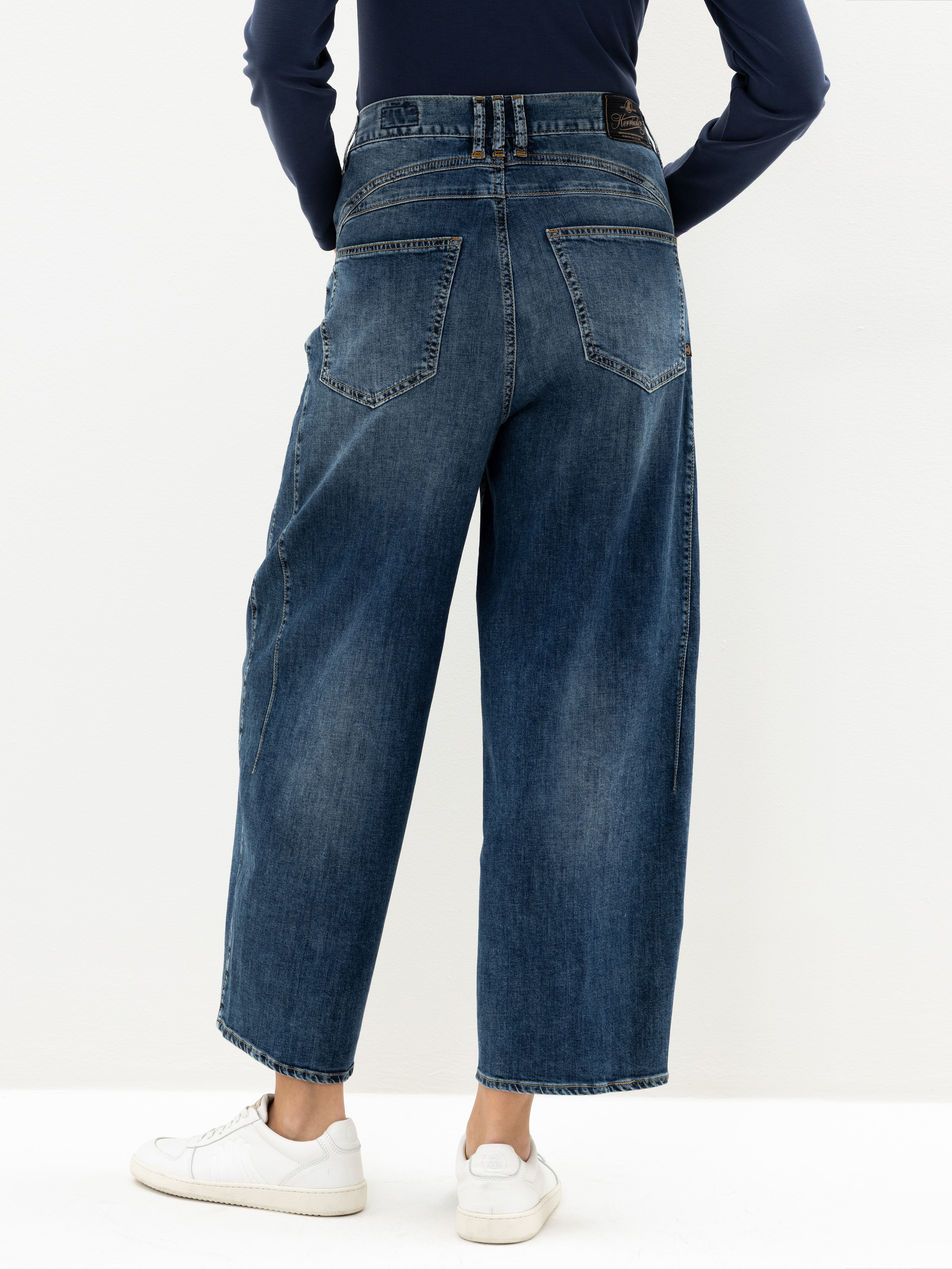 Eine Person, die weit geschnittene blaue Jeans, ein marineblaues langärmeliges Oberteil und weiße Turnschuhe trägt, steht vor einem schlichten weißen Hintergrund, von hinten gesehen.