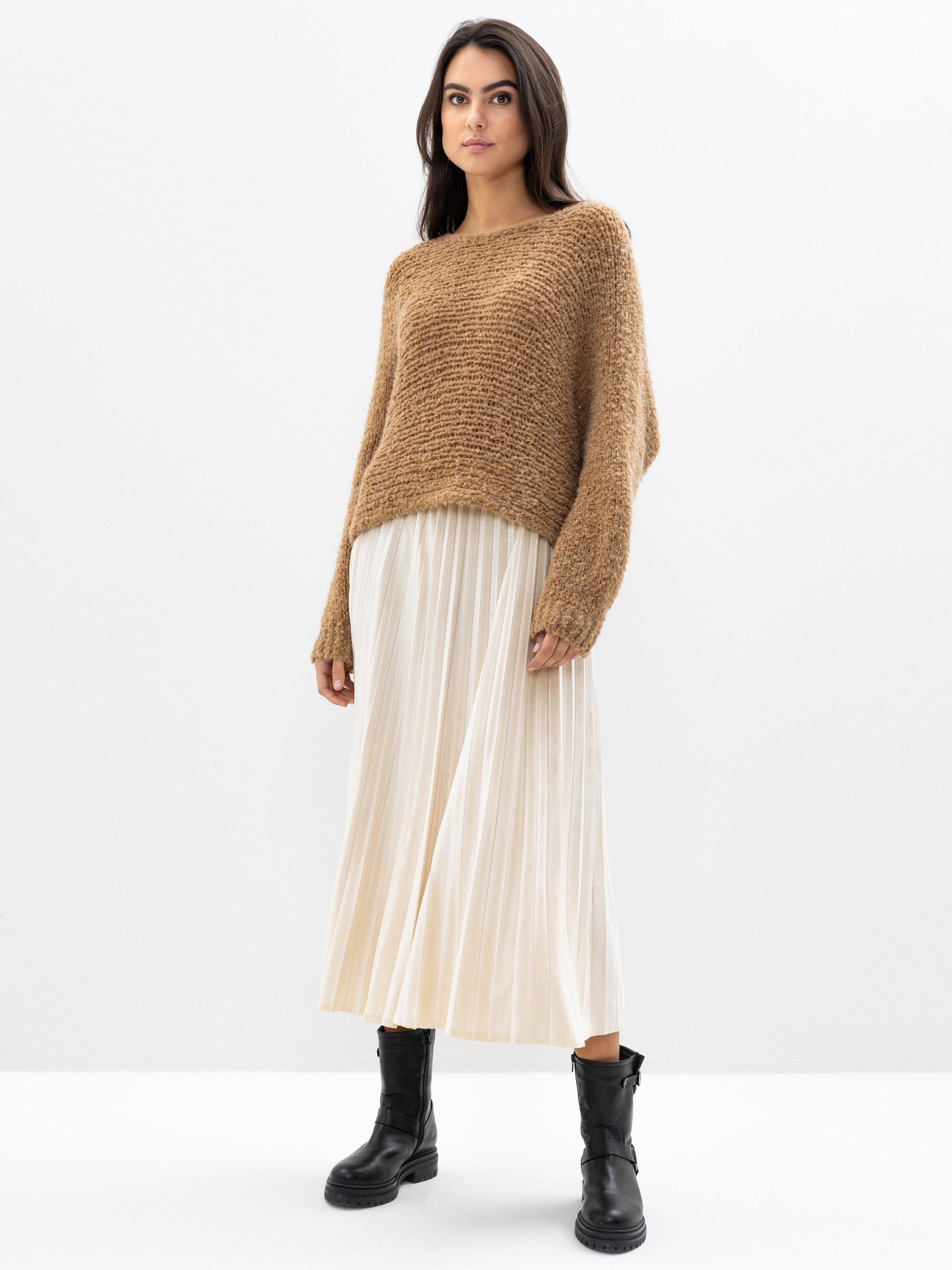 Eine Frau steht vor einem schlichten weißen Hintergrund, trägt einen lockeren braunen Strickpulli, einen cremefarbenen Falten-Midirock und schwarze Stiefeletten und blickt mit einem entspannten Gesichtsausdruck leicht zur Seite.