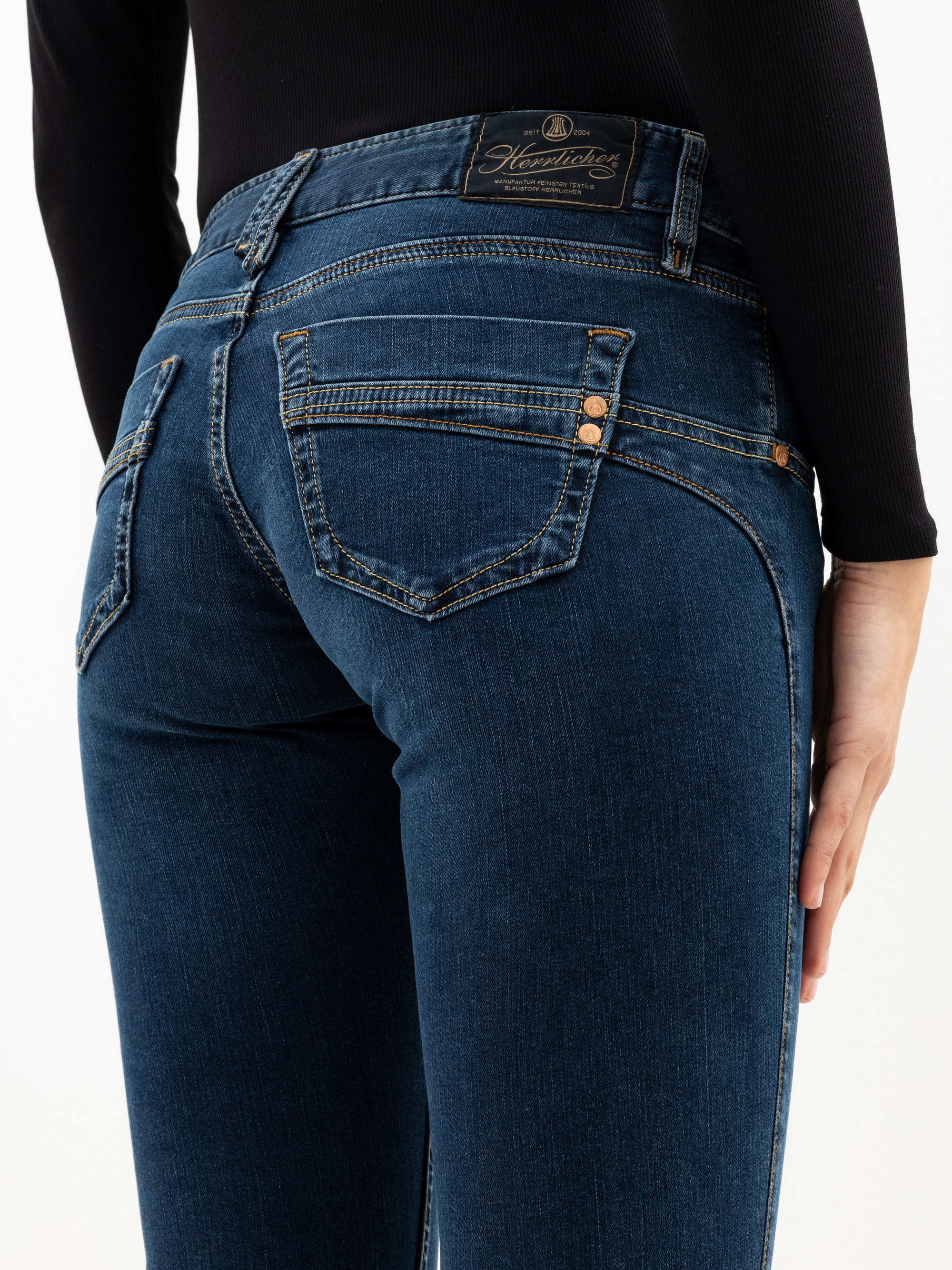 Eine Person trägt eine dunkelblaue Jeans und ein schwarzes langärmeliges Oberteil, von hinten gesehen. Die Jeans hat sichtbare Nähte, zwei Gesäßtaschen und ein Markenetikett am Bund.