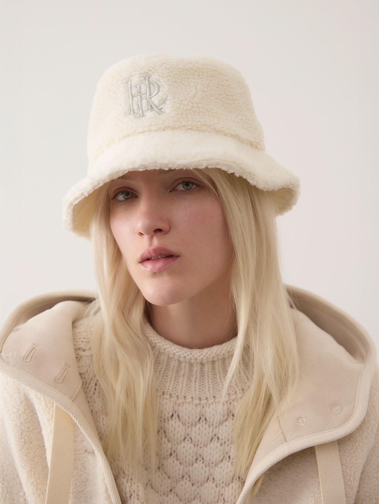 Eine Frau mit langen blonden Haaren trägt eine cremefarbene Sherpa-Mütze mit aufgestickten Initialen, einen strukturierten Strickpullover und eine passende Jacke vor einem schlichten hellen Hintergrund.