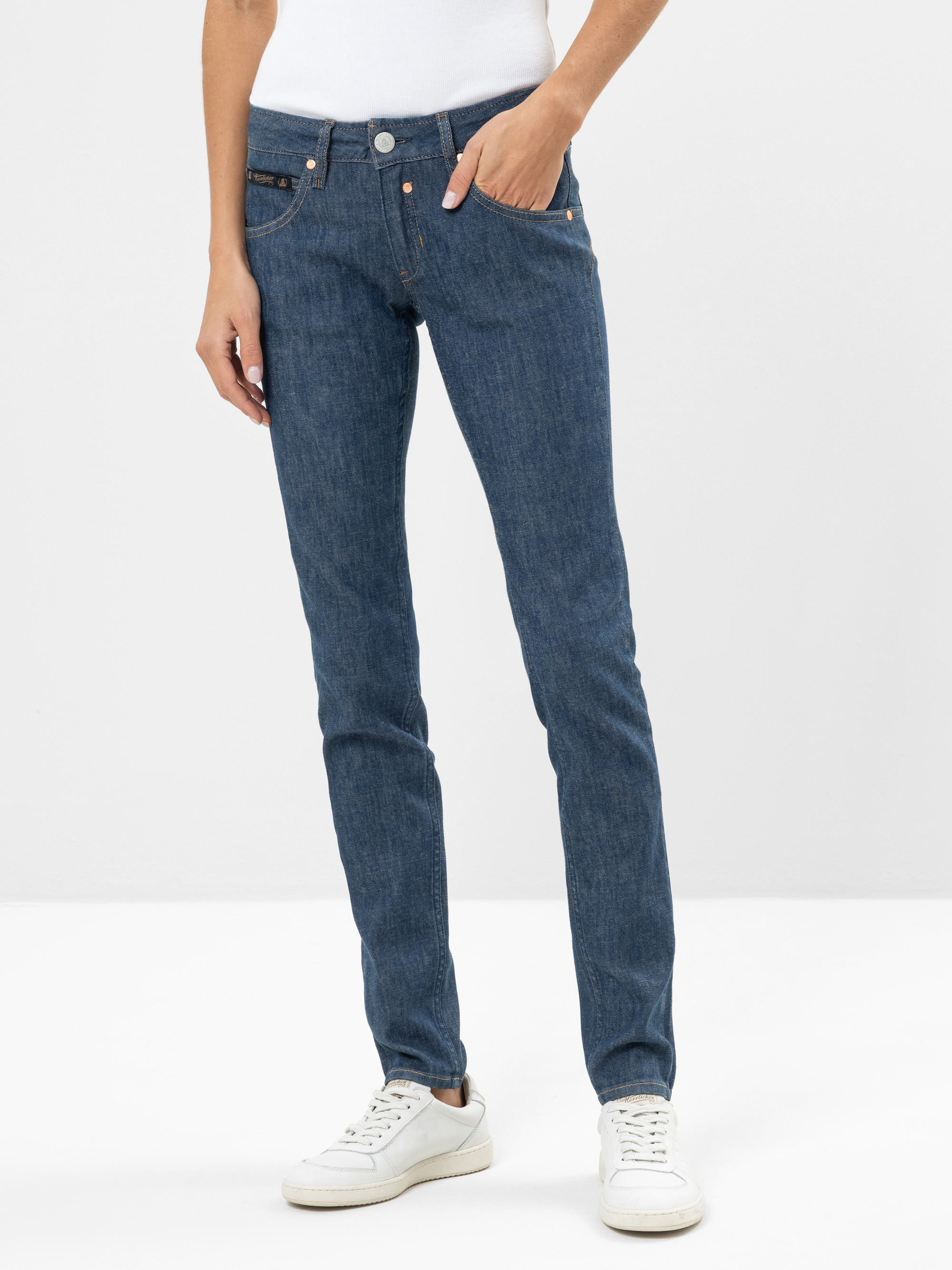 Eine Person in blauen Jeans und weißen Turnschuhen steht vor einem schlichten weißen Hintergrund, eine Hand in der Hosentasche, den anderen Arm locker an der Seite.