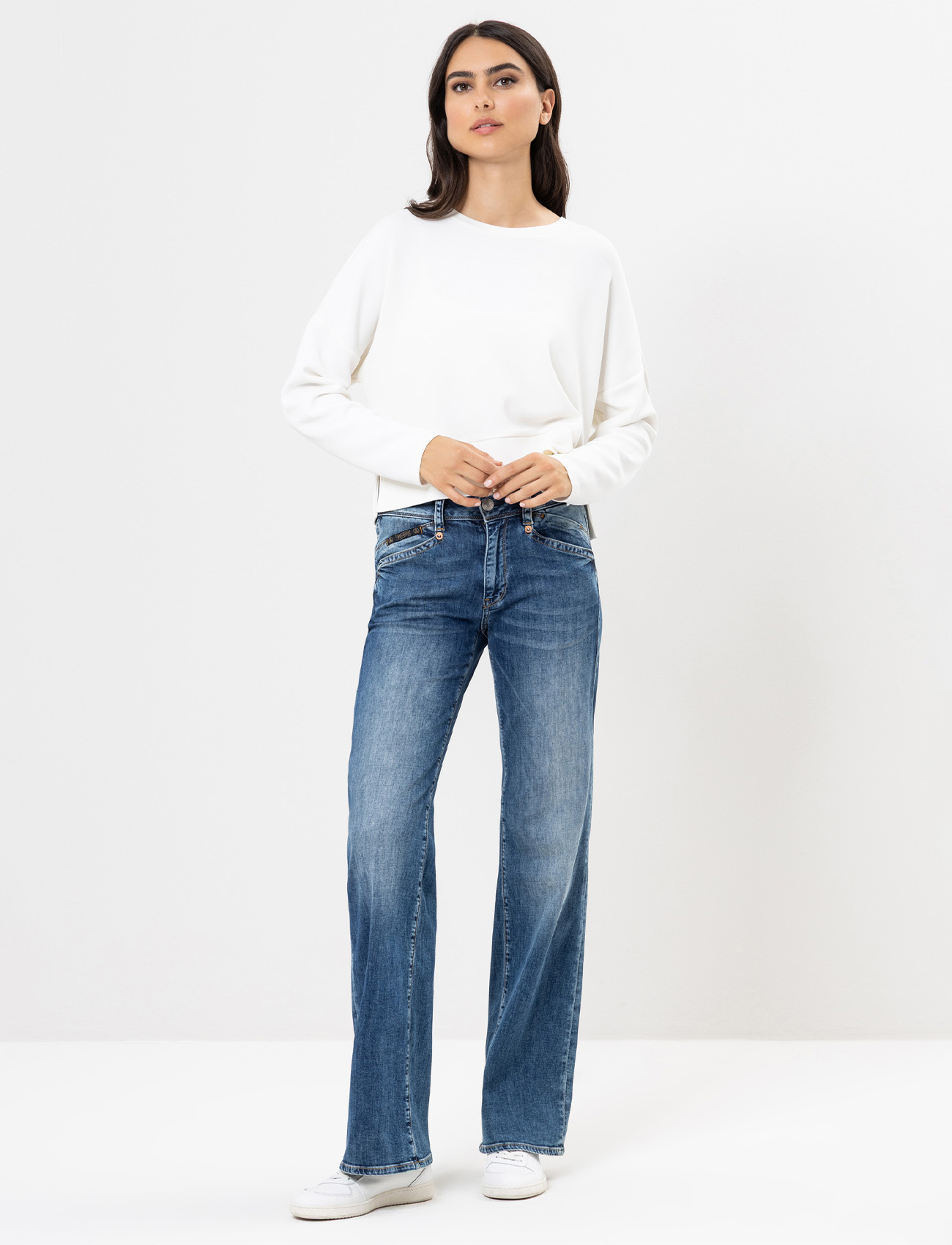 Eine Frau steht vor einem schlichten weißen Hintergrund, trägt ein weißes langärmeliges Oberteil, blaue Jeans und weiße Turnschuhe und hat die Hände in den Taschen ihrer Jeans.