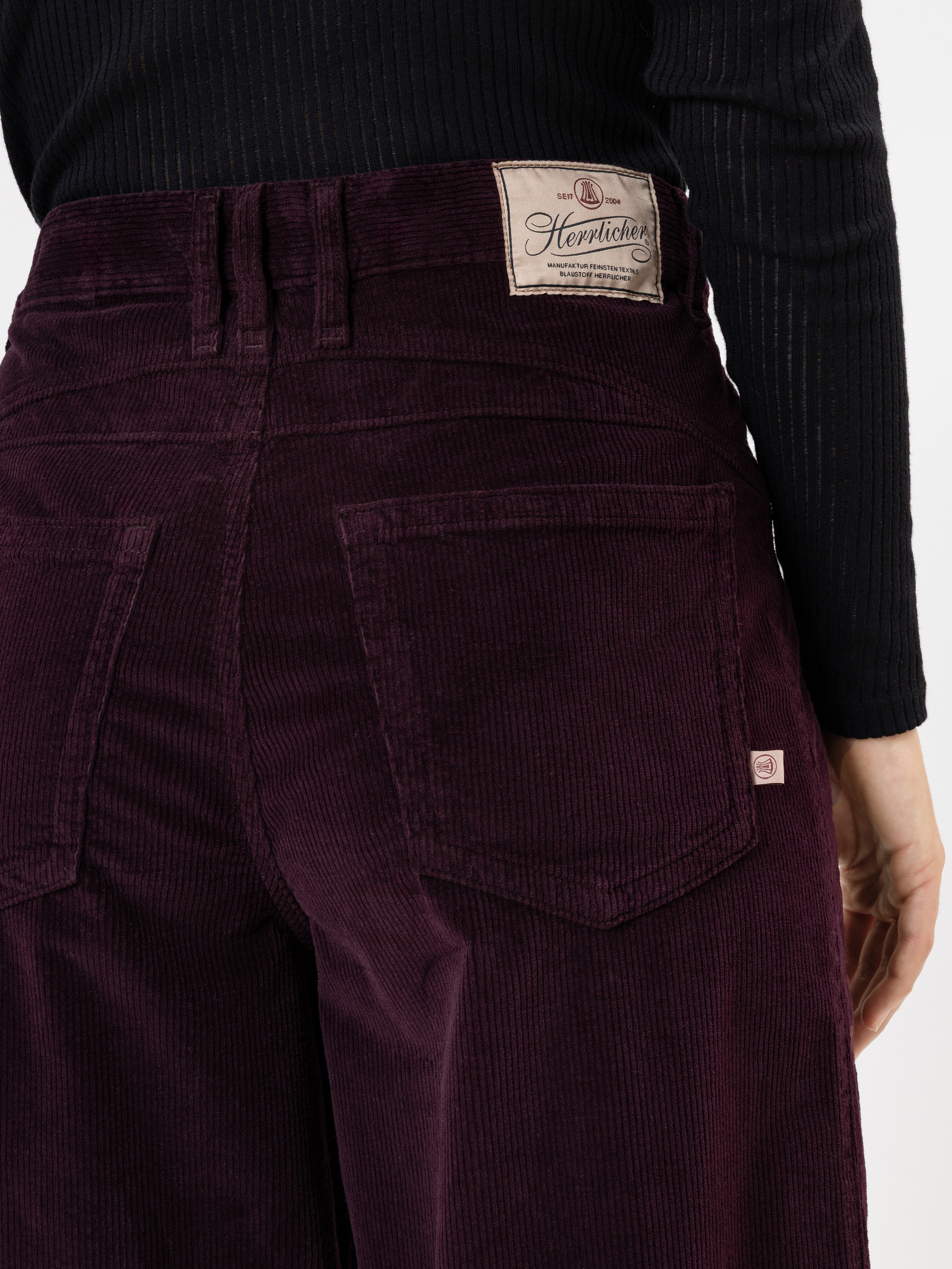Eine Person, die eine dunkelviolette Cordhose mit großen Gesäßtaschen und einem hellbraunen Herrlicher-Markenetikett trägt, dazu ein schwarzes langärmeliges Rippenoberteil.