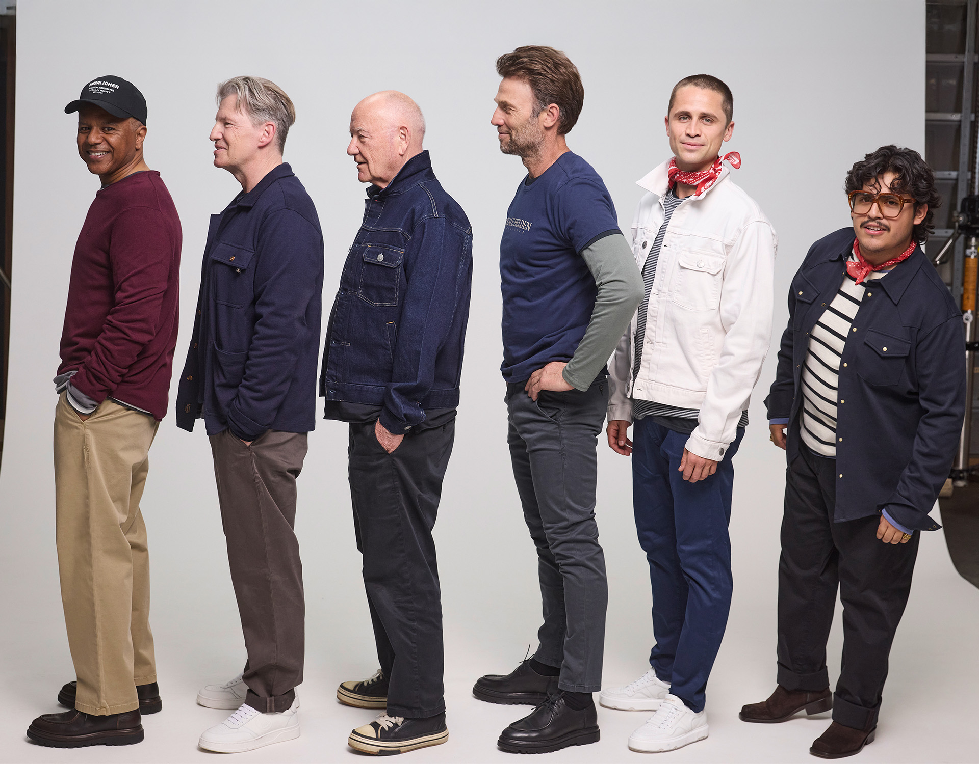 Sechs Männer unterschiedlichen Alters und unterschiedlicher Statur stehen in einer Reihe, nach links gewandt, vor einem schlichten Studiohintergrund. Sie tragen legere Kleidung wie Jacken, Jeans und Turnschuhe und haben einen entspannten und freundlichen